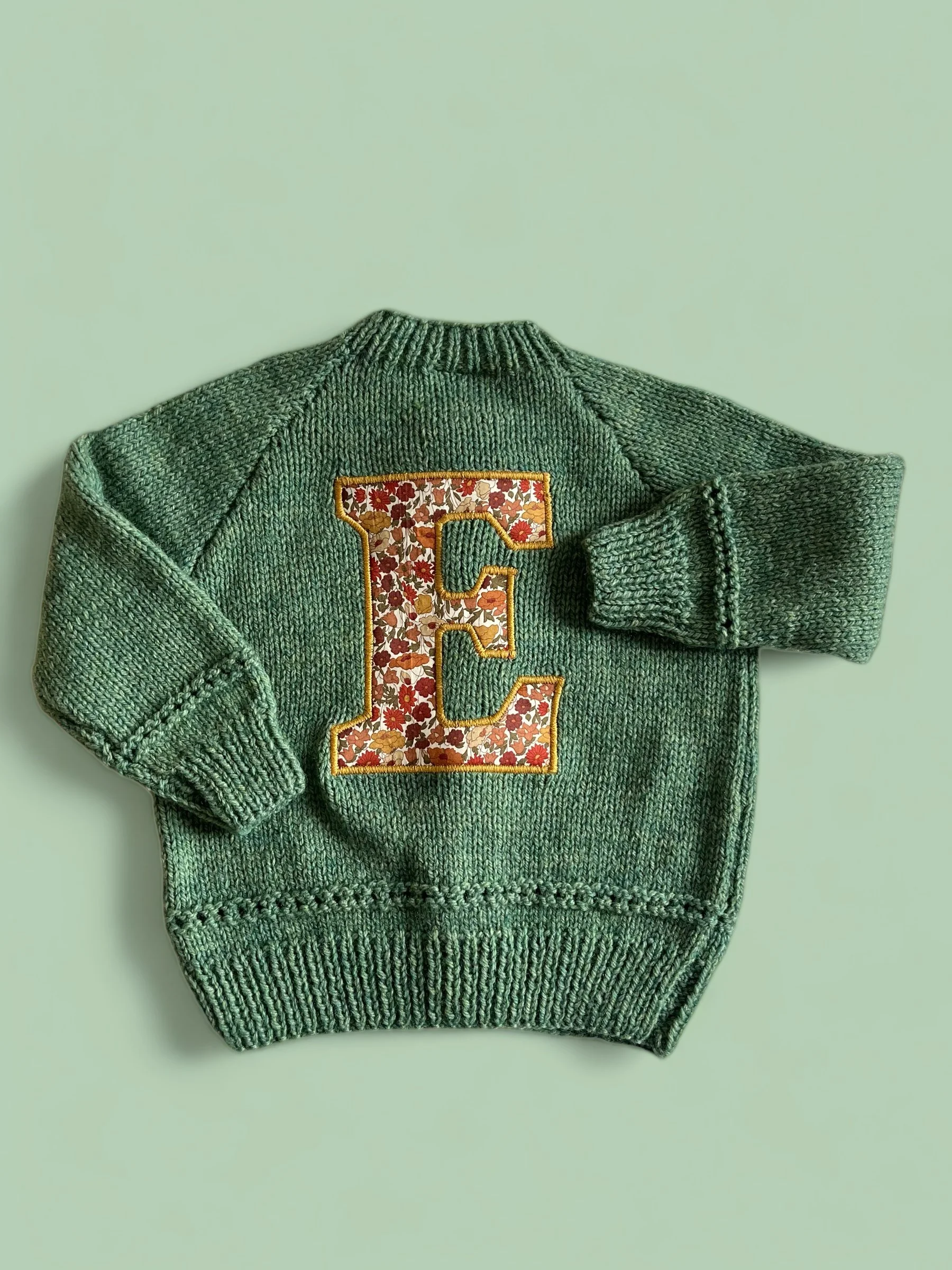 Green knitted sweater with a large letter 'E' patch on the front, decorated with a floral pattern.