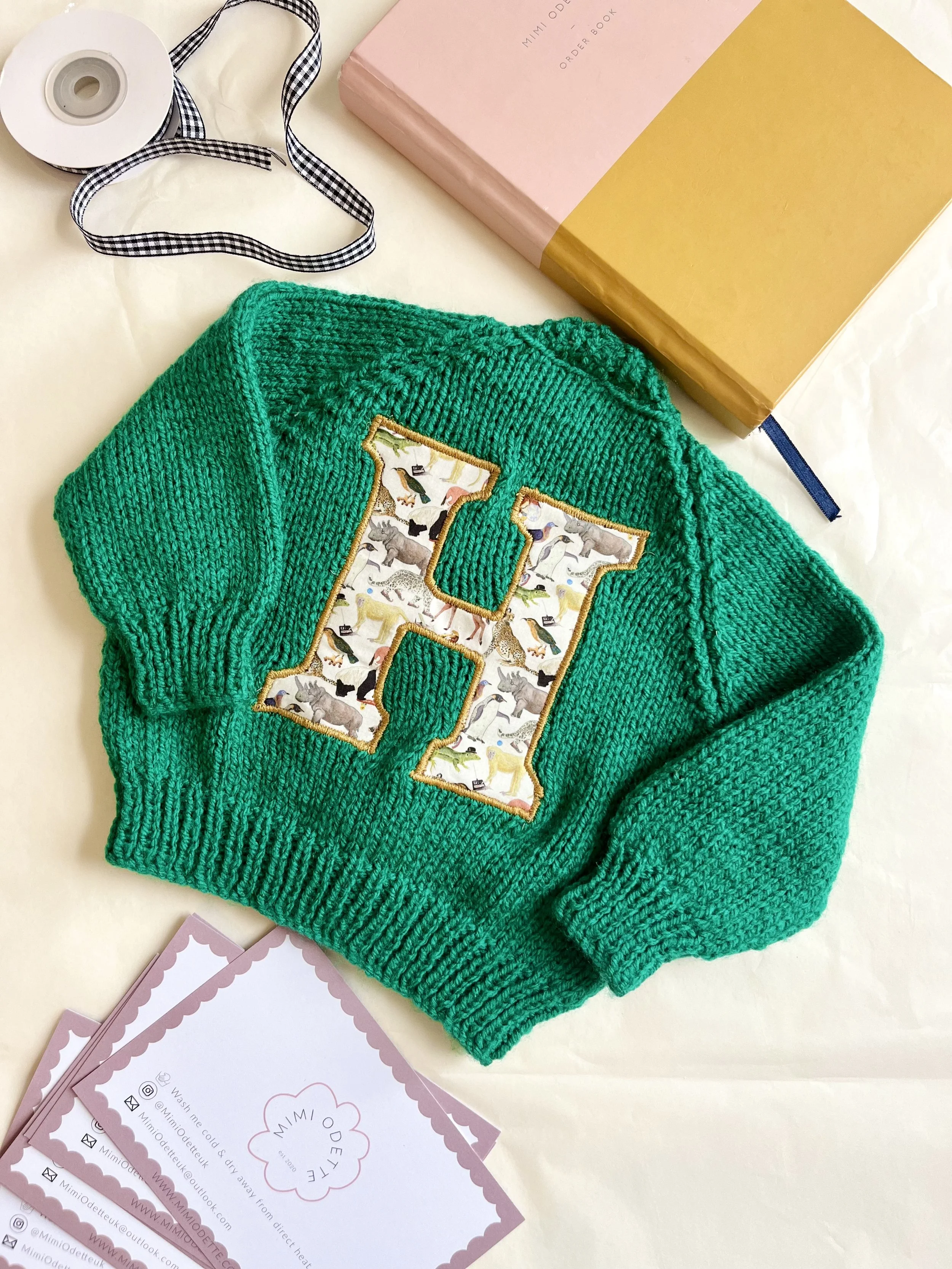 A green knitted sweater with a large embroidered letter 'H' on the front, surrounded by a fabric pattern of various animals. The sweater is on a flat surface alongside business cards, a pink and gold notebook, a roll of tape, a black-and-white checke