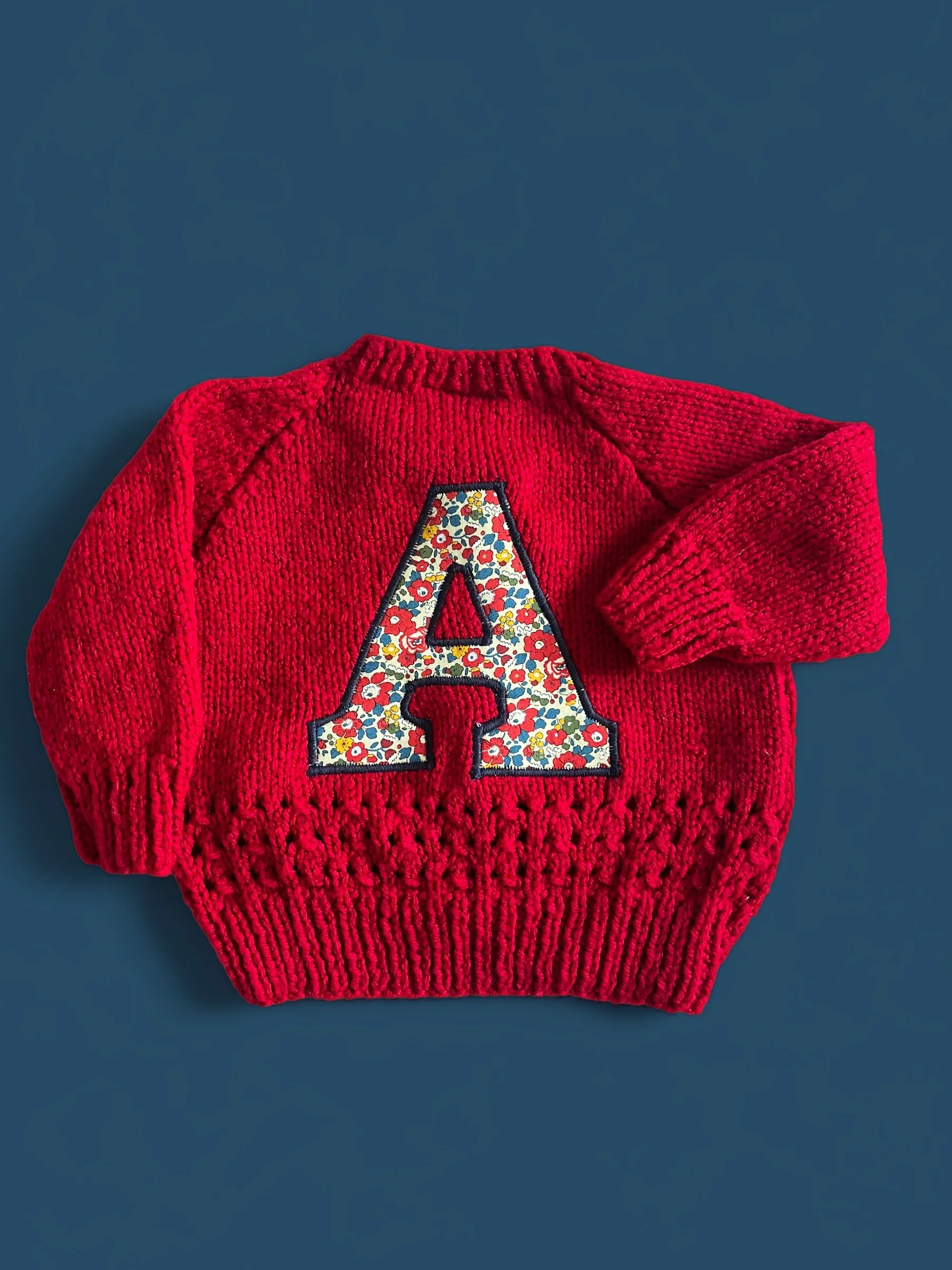 Red knitted sweater with a large embroidered letter 'A' decorated with a colorful floral pattern, placed on a dark blue surface.