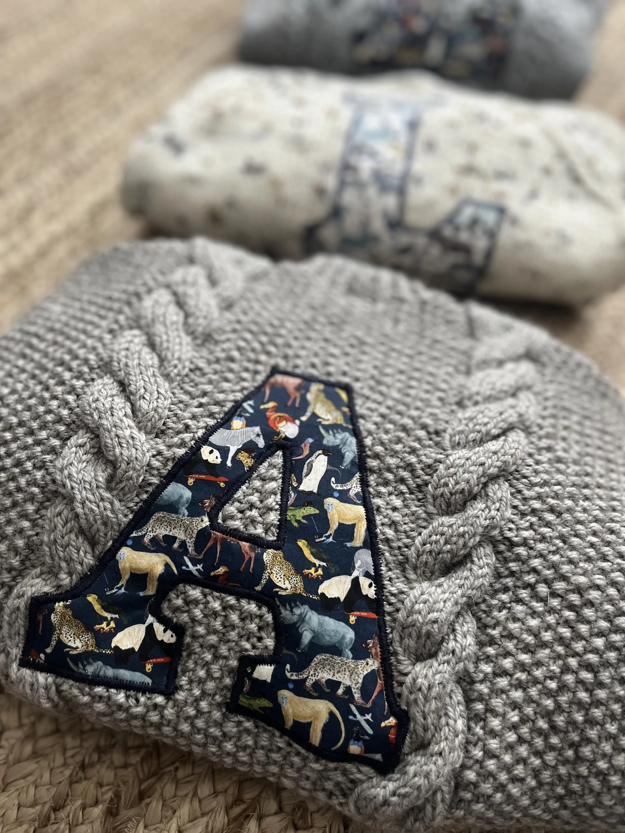 Close-up of a gray knitted sweater featuring a large letter 'A' with an animal print design, placed on a textured surface with more sweaters in the background.