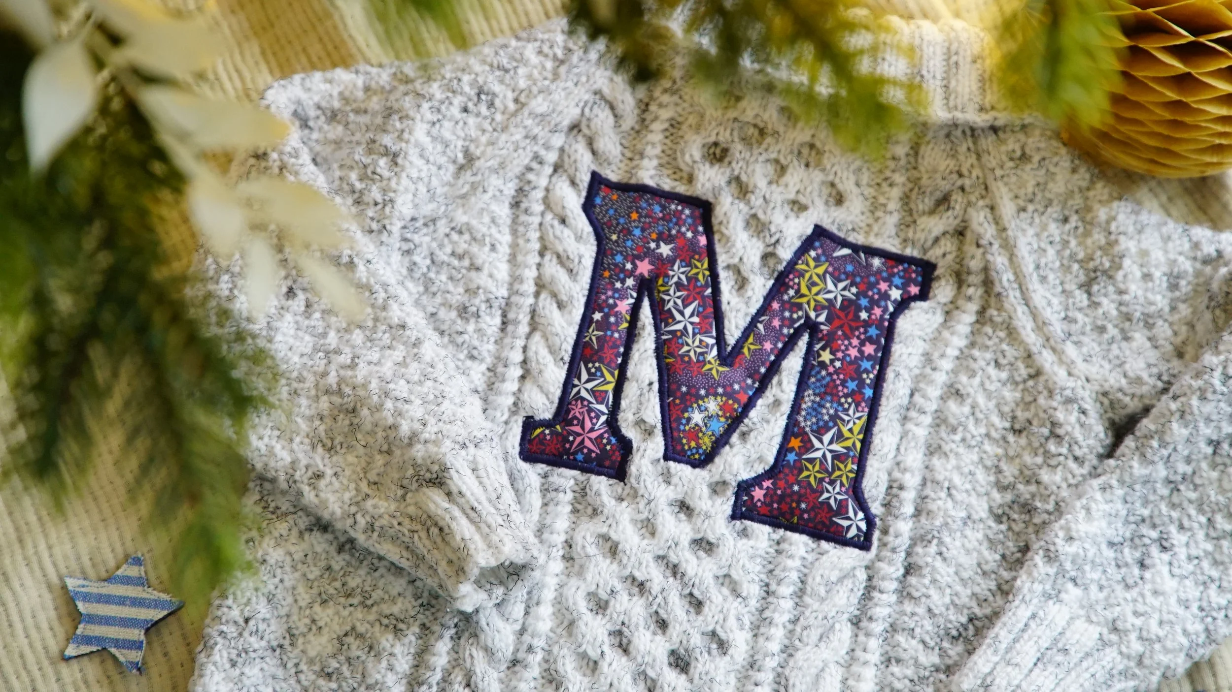 Gray knit sweater with colorful letter 'M' appliqué, surrounded by green branches and decorative paper star.