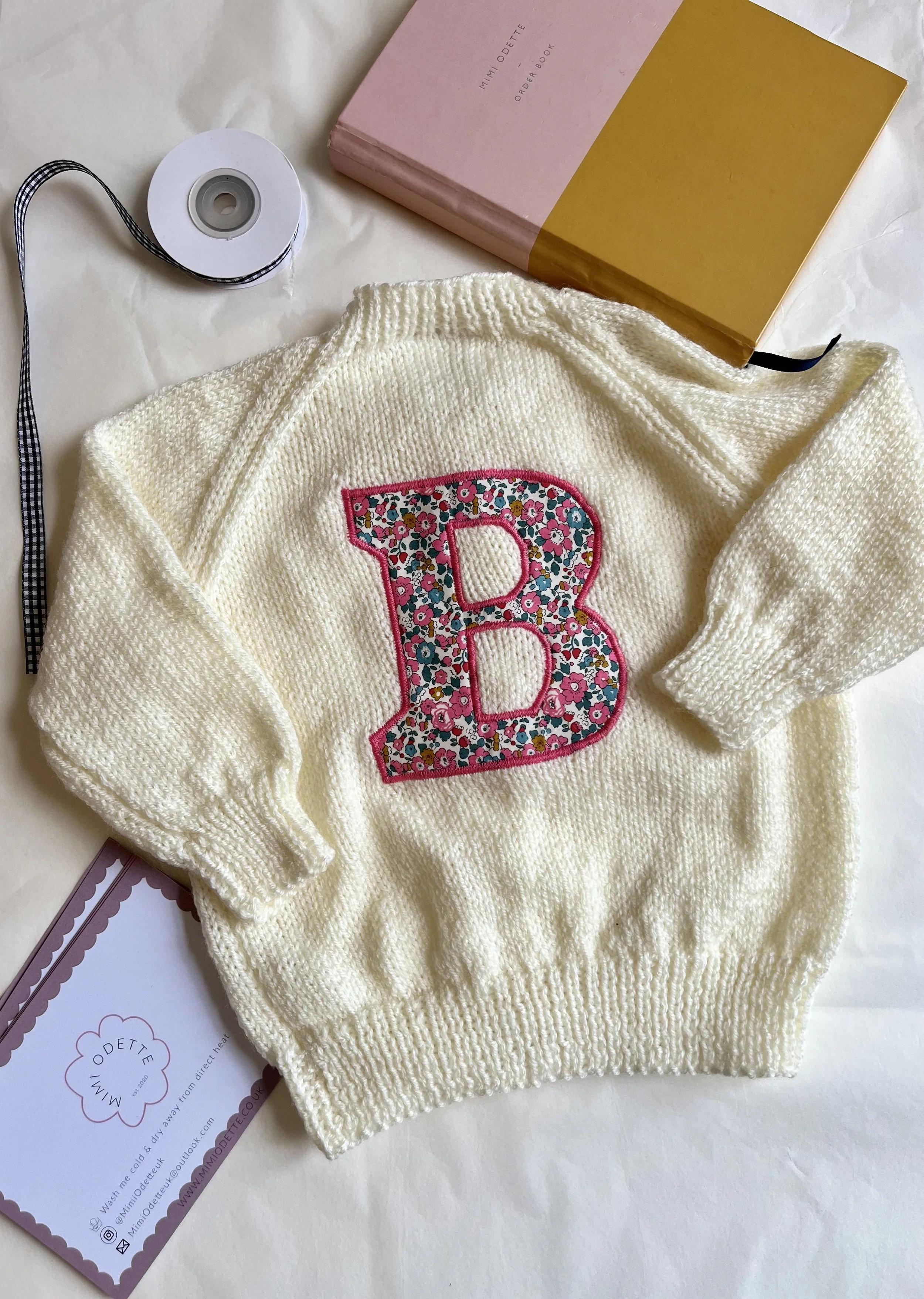 Cream knitted sweater with floral 'B' applique, pink and gold book, ribbon spool, and a card.