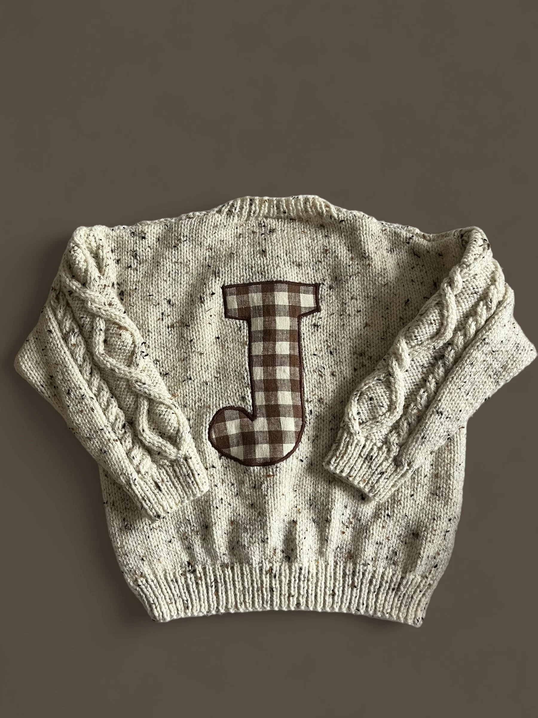 Cream-colored knit sweater with brown and white checkered letter 'J' in the center and braided cable knit design on the sleeves.