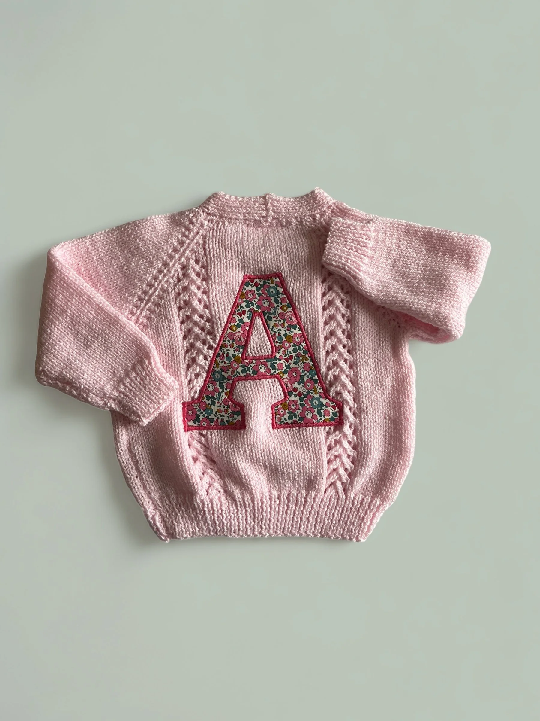 Pink knitted sweater with a large floral embroidered letter A in the center.
