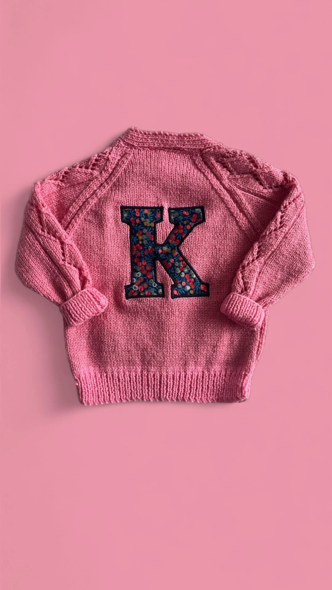 Pink knit sweater with a floral letter K on the back, set against a pink background.
