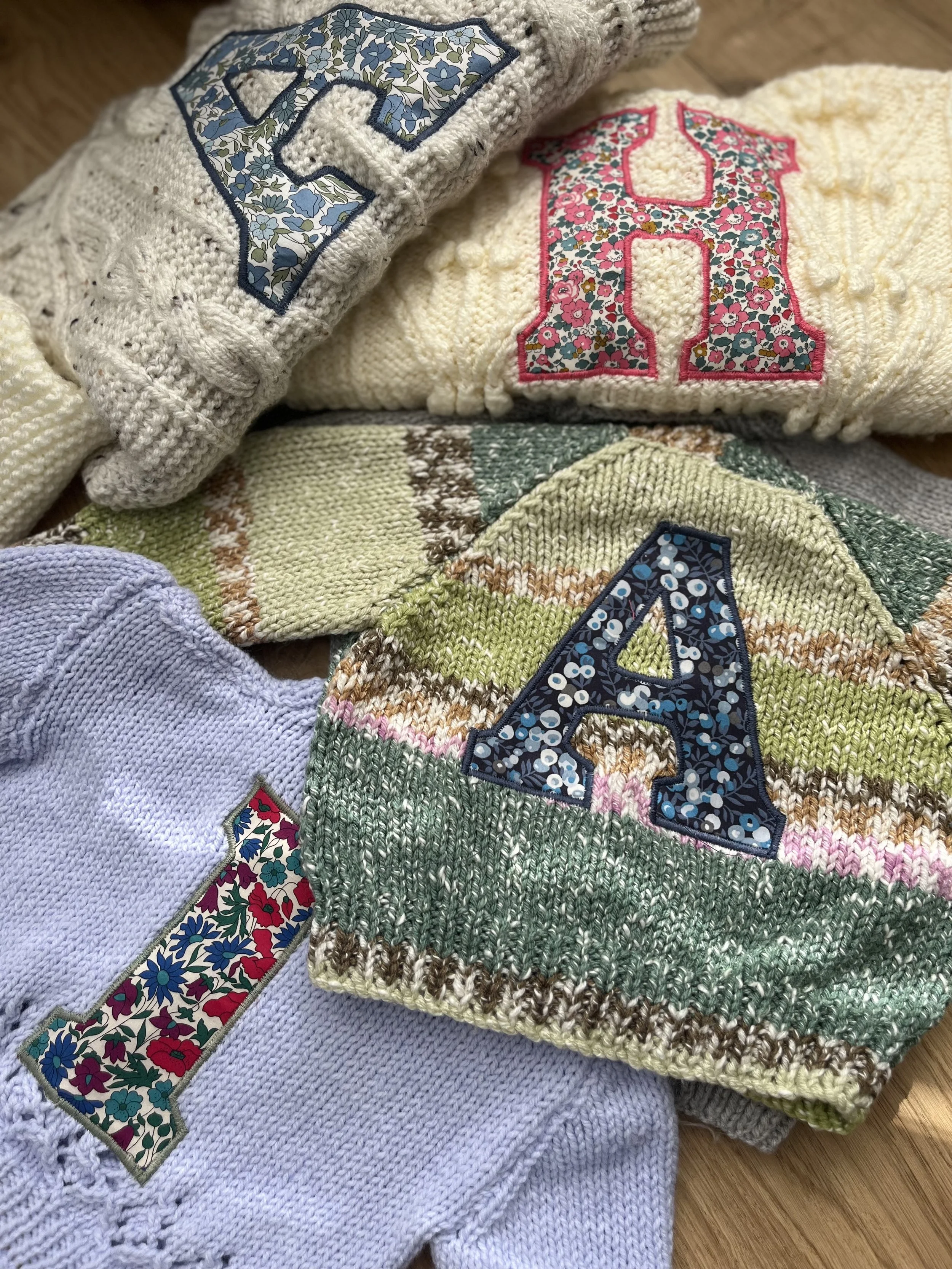 A collection of knitted sweaters featuring large floral-patterned letters "A" and "H" on a wooden surface.