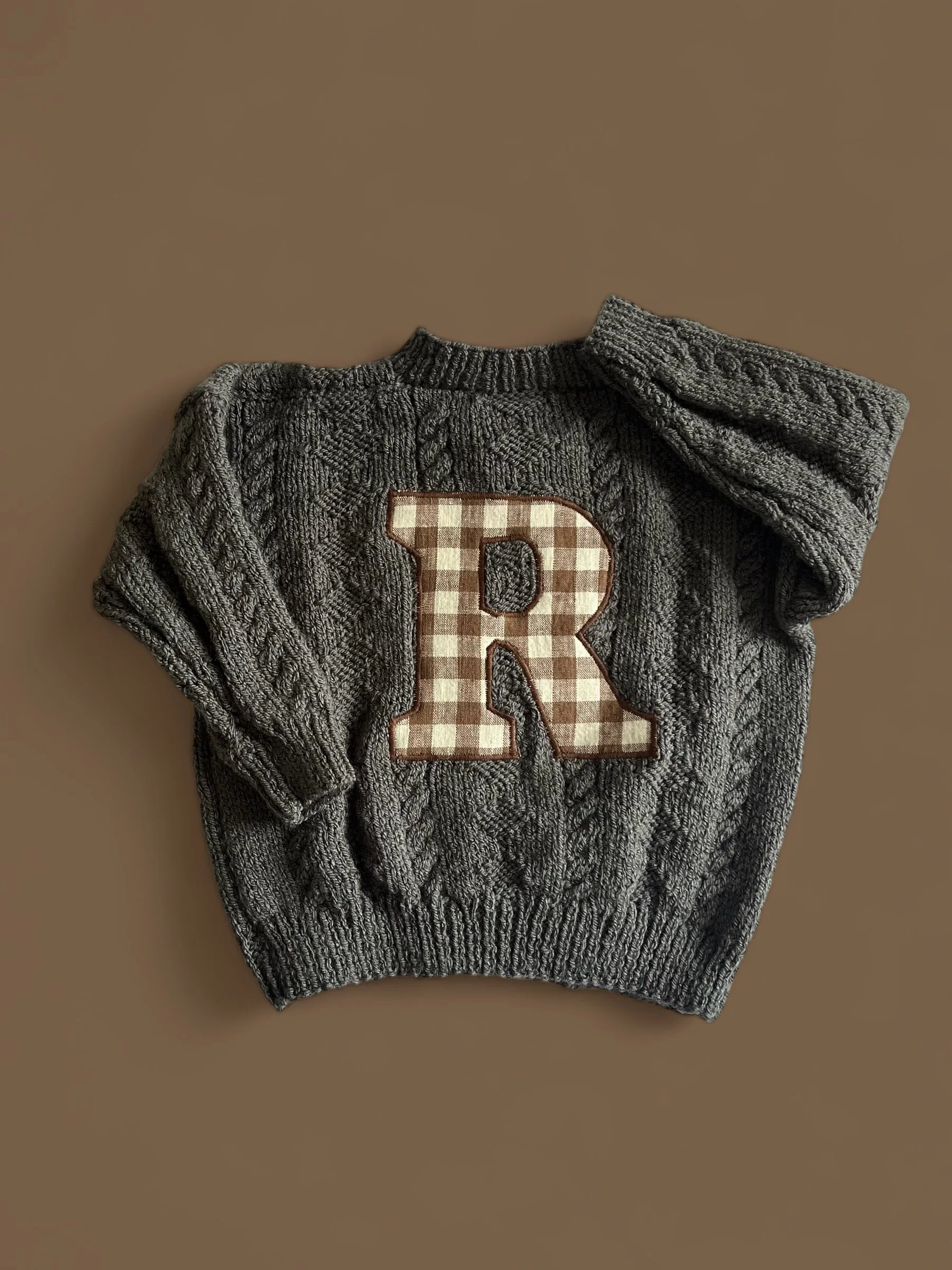 A gray knit sweater with a large checkered letter R patch on the front, laid flat on a brown surface.