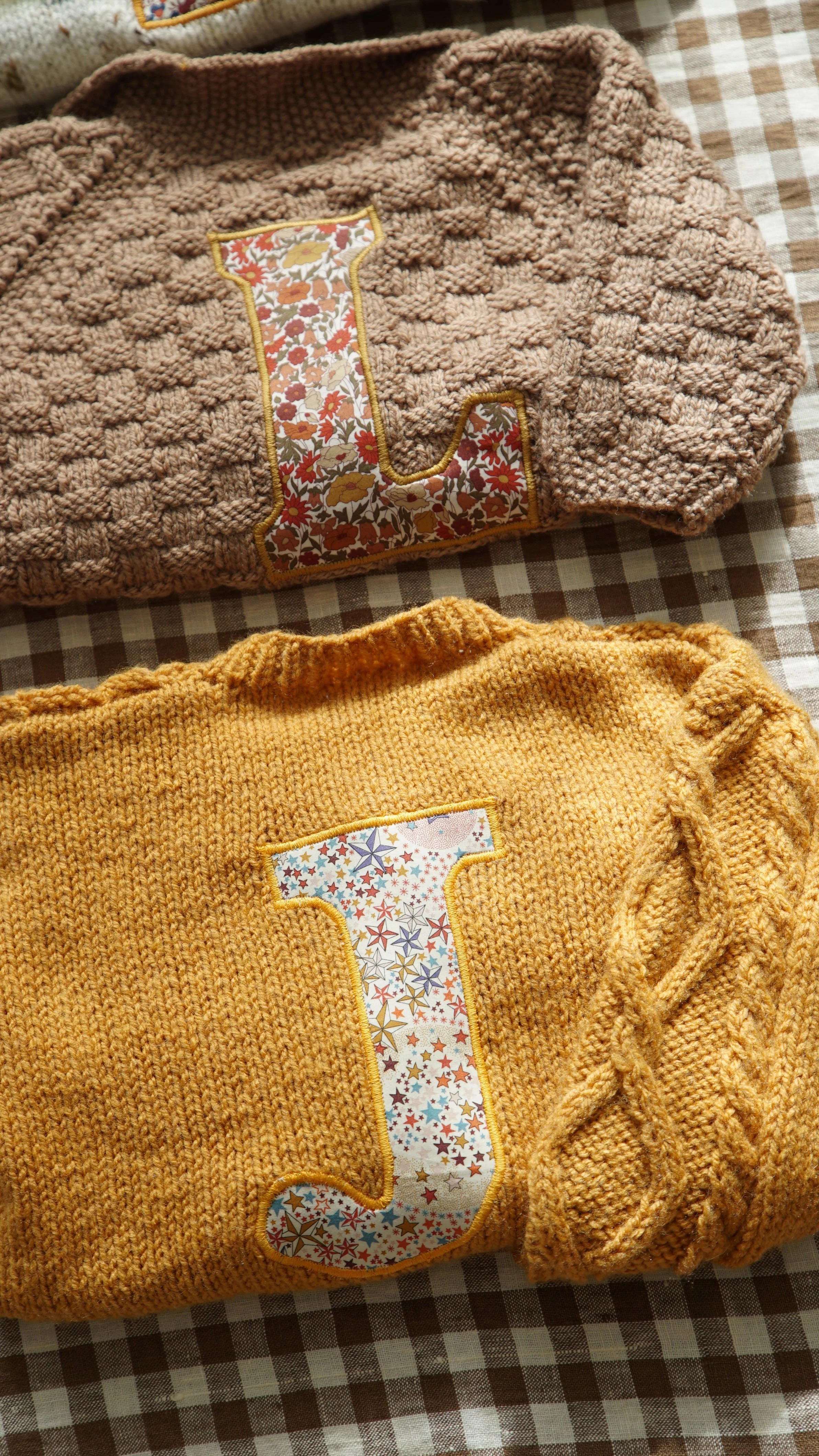 Two knitted sweaters with fabric applique letters, "L" on a brown sweater and "J" on a mustard-yellow sweater, displayed on a checkered surface.