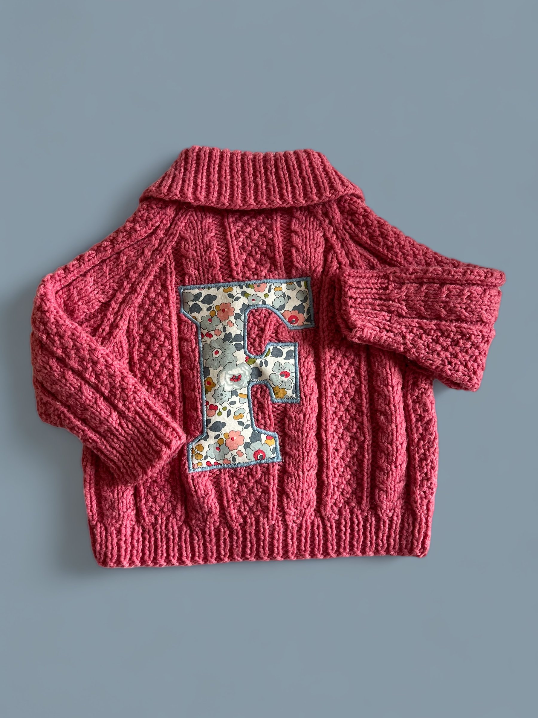 Pink knitted cardigan sweater with a floral embroidered letter 'E' on the front, placed on a gray background.