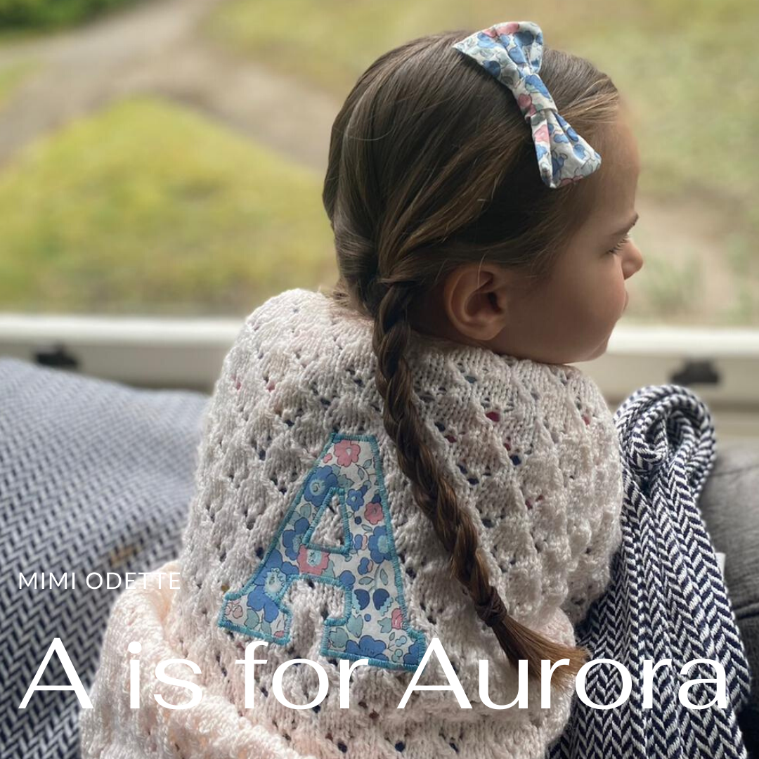 Young girl with braids and bow in her hair, wearing a crocheted cardigan with a floral letter 'A' patch, sitting by a window.