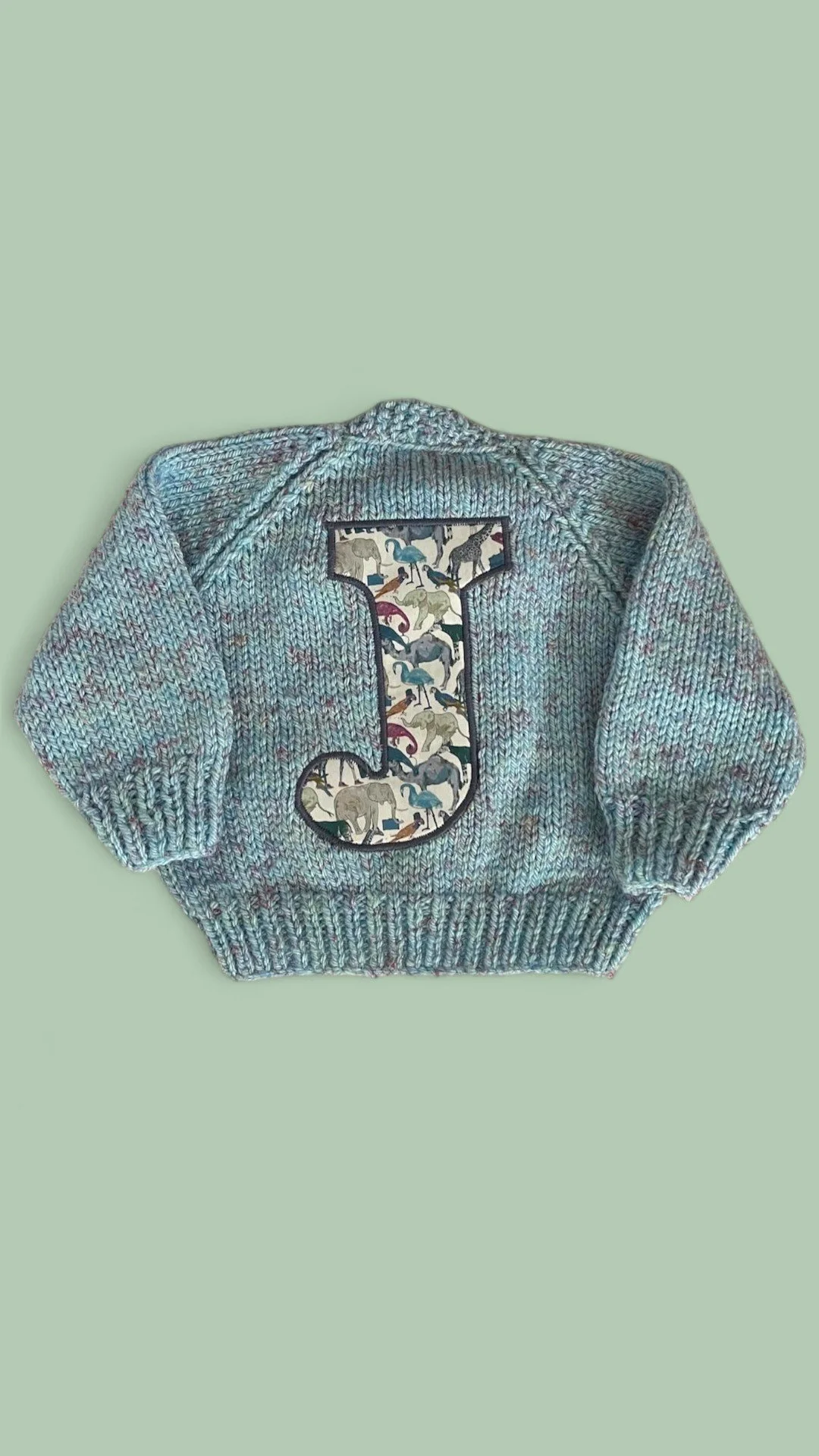A knitted baby sweater in blue tones with a large patch letter 'J' featuring a zoo animal pattern.