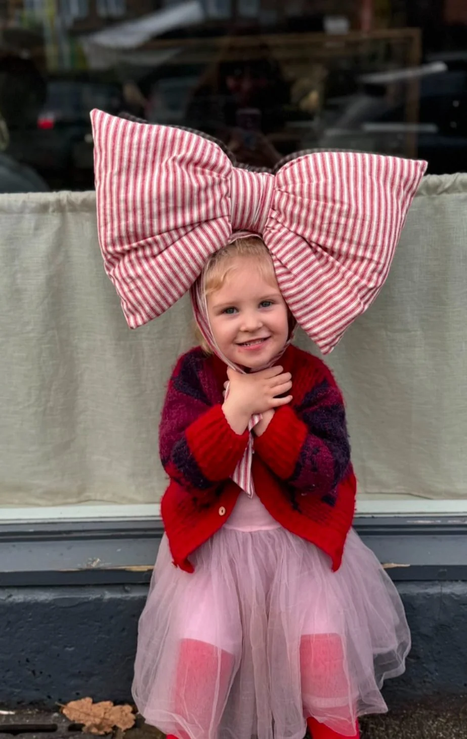 Child wearing a large red-and-white striped bow on their head, dressed in a red sweater and pink tulle skirt, smiling with hands clasped.