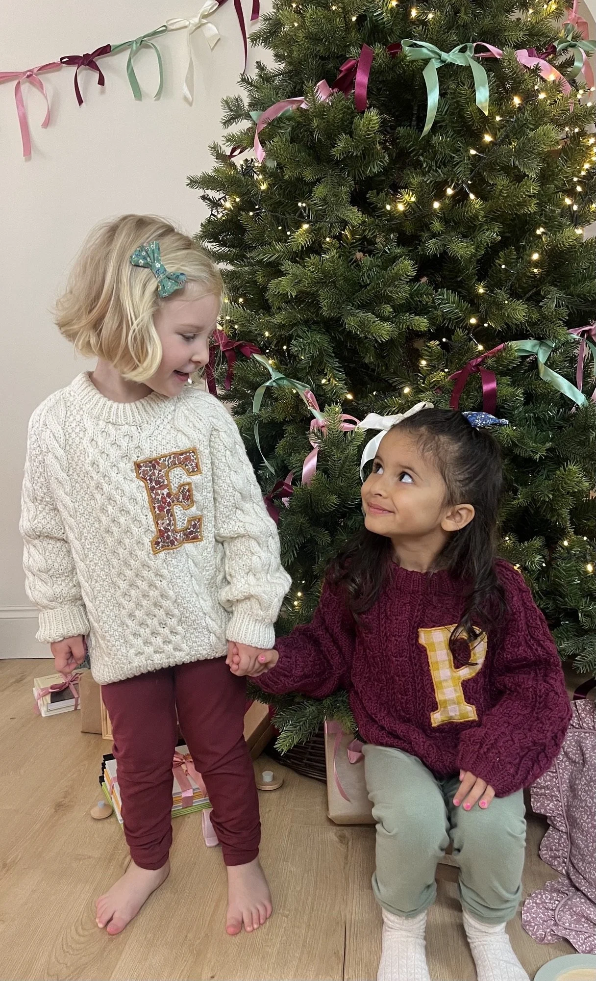 Two children wearing monogrammed sweaters holding hands in front of a decorated Christmas tree with lights and ribbons.
