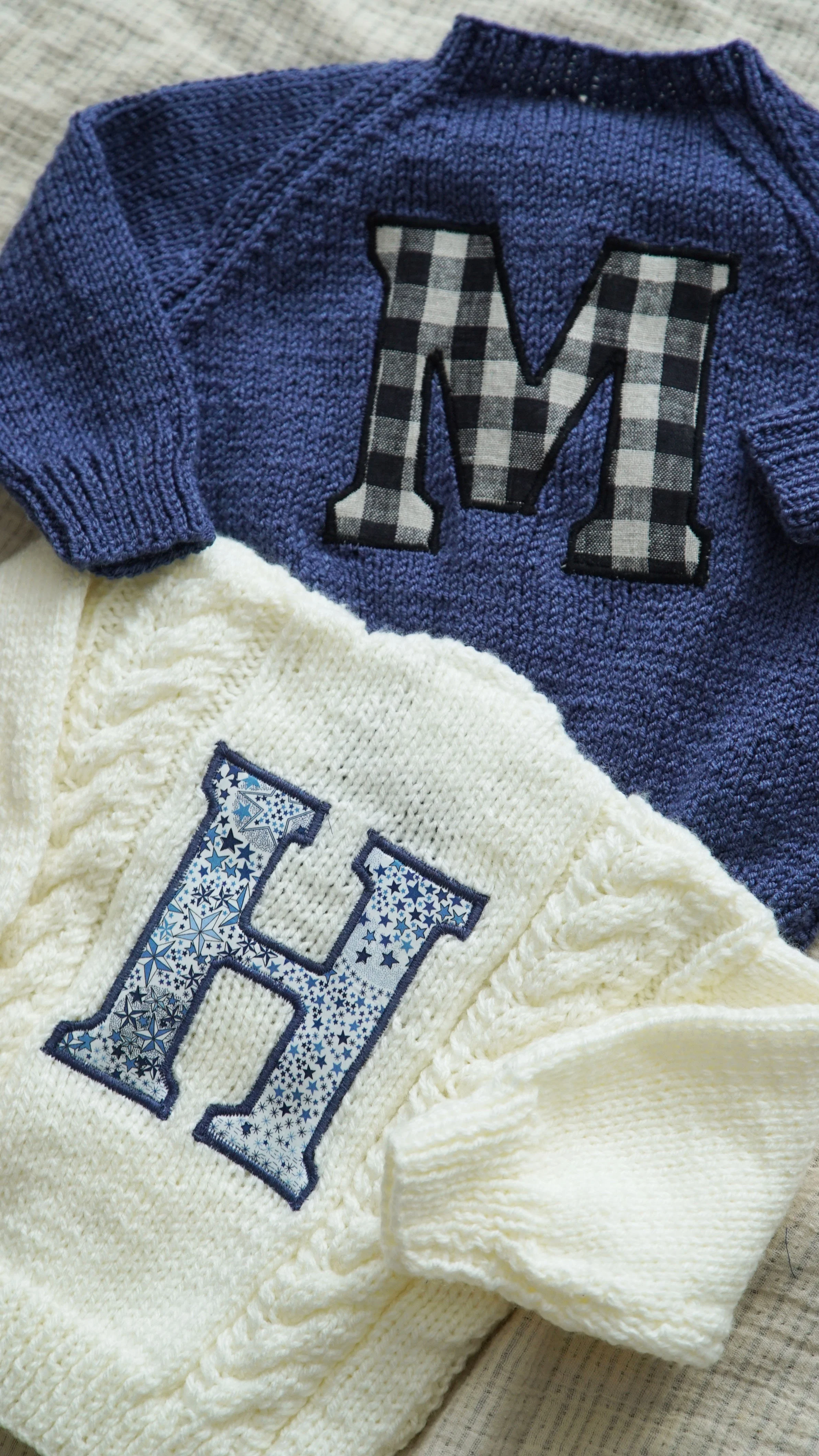 Two knitted sweaters, one blue with a checkered 'M', and one white with a star-patterned 'H'. The sweaters are laid on a textured fabric surface.