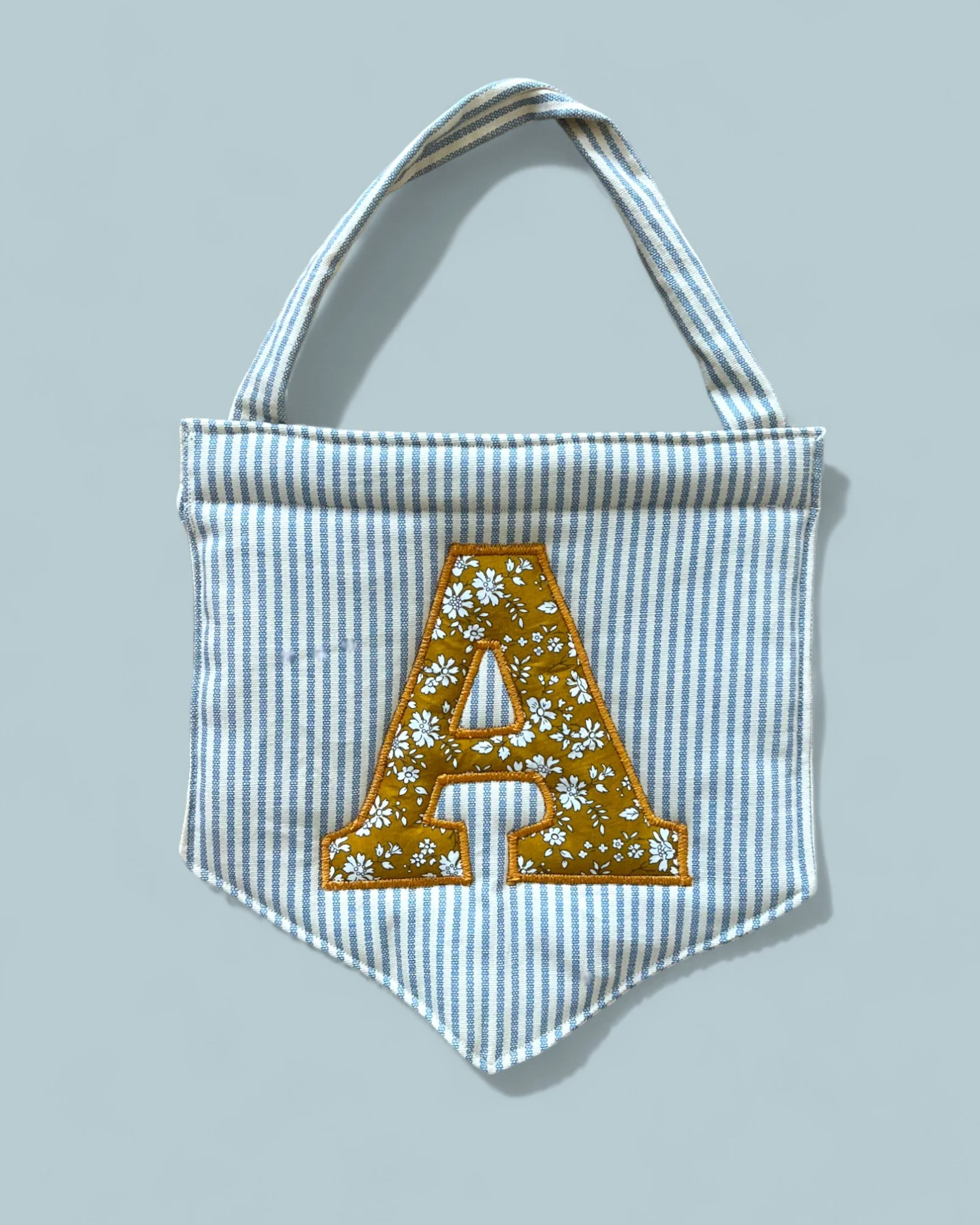 A fabric tote bag with blue and white vertical stripes, featuring a large yellow letter 'A' with a white floral pattern in the center.