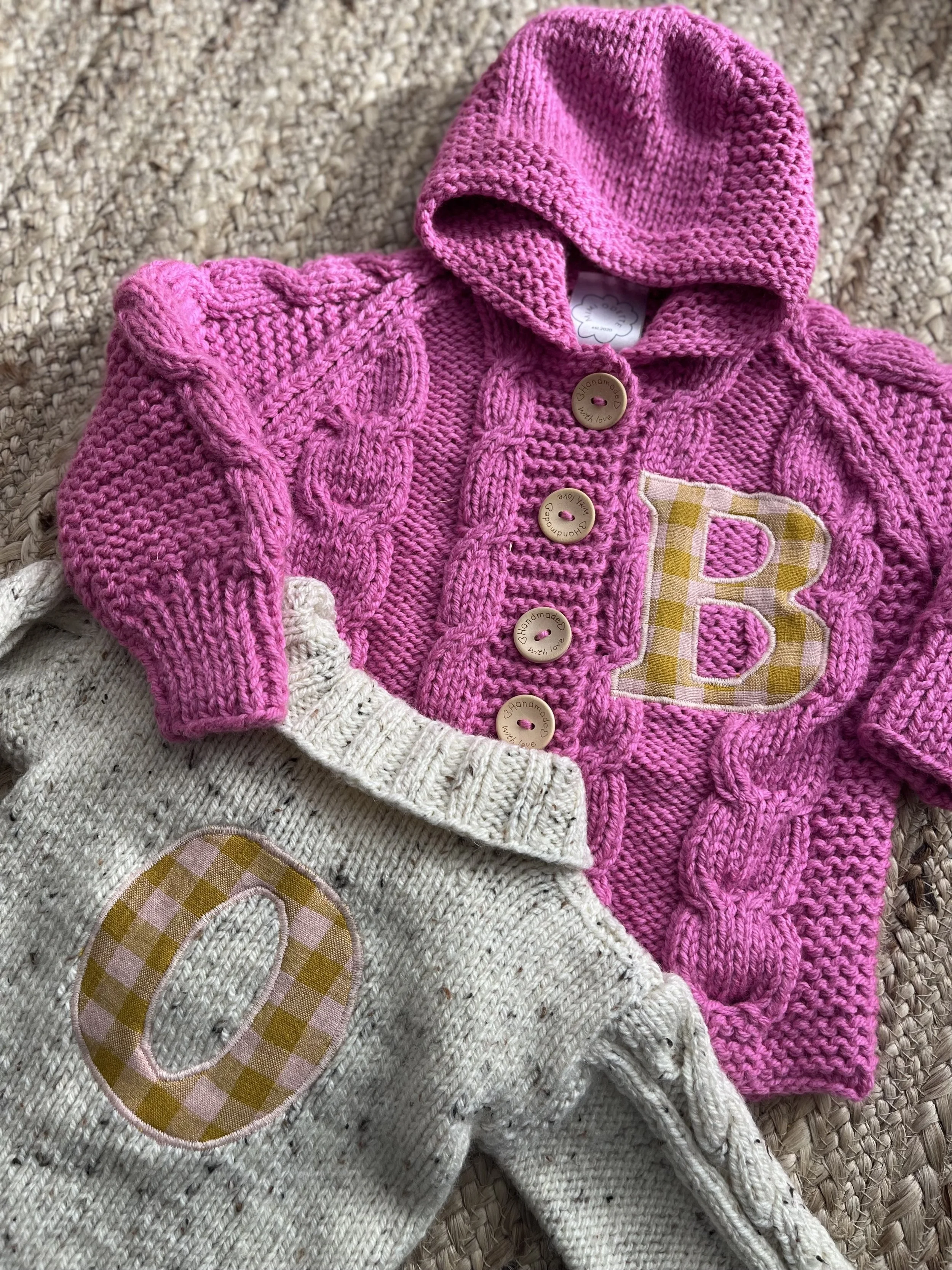 Pink knitted hoodie with large buttons and letter 'B,' and a gray knitted sweater with letter 'O' on textured background.