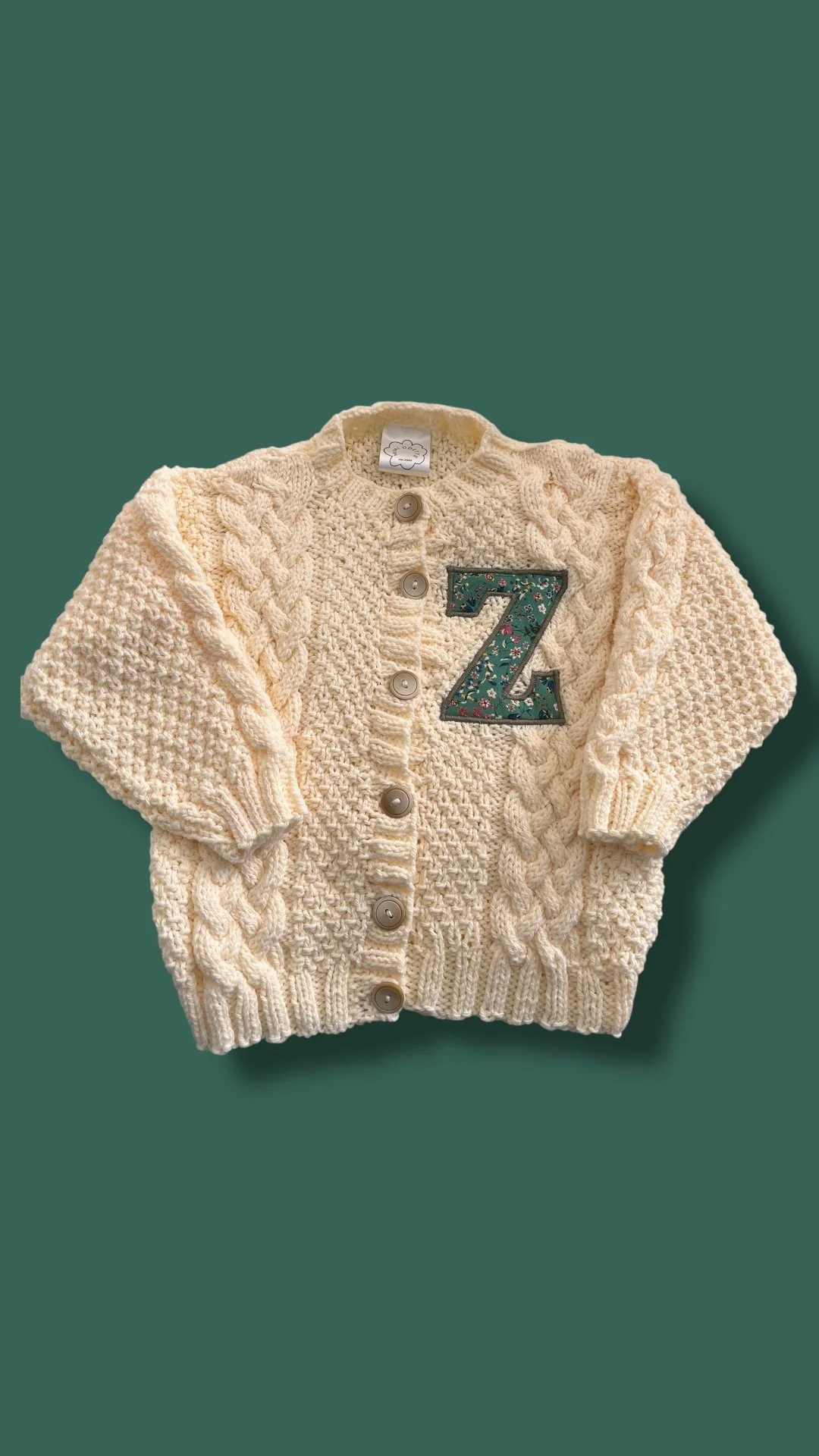 Cream-colored knitted cardigan with cable knit pattern, buttons down the front, and a decorative patch of the letter 'Z' on the left chest, laid flat on a green background.