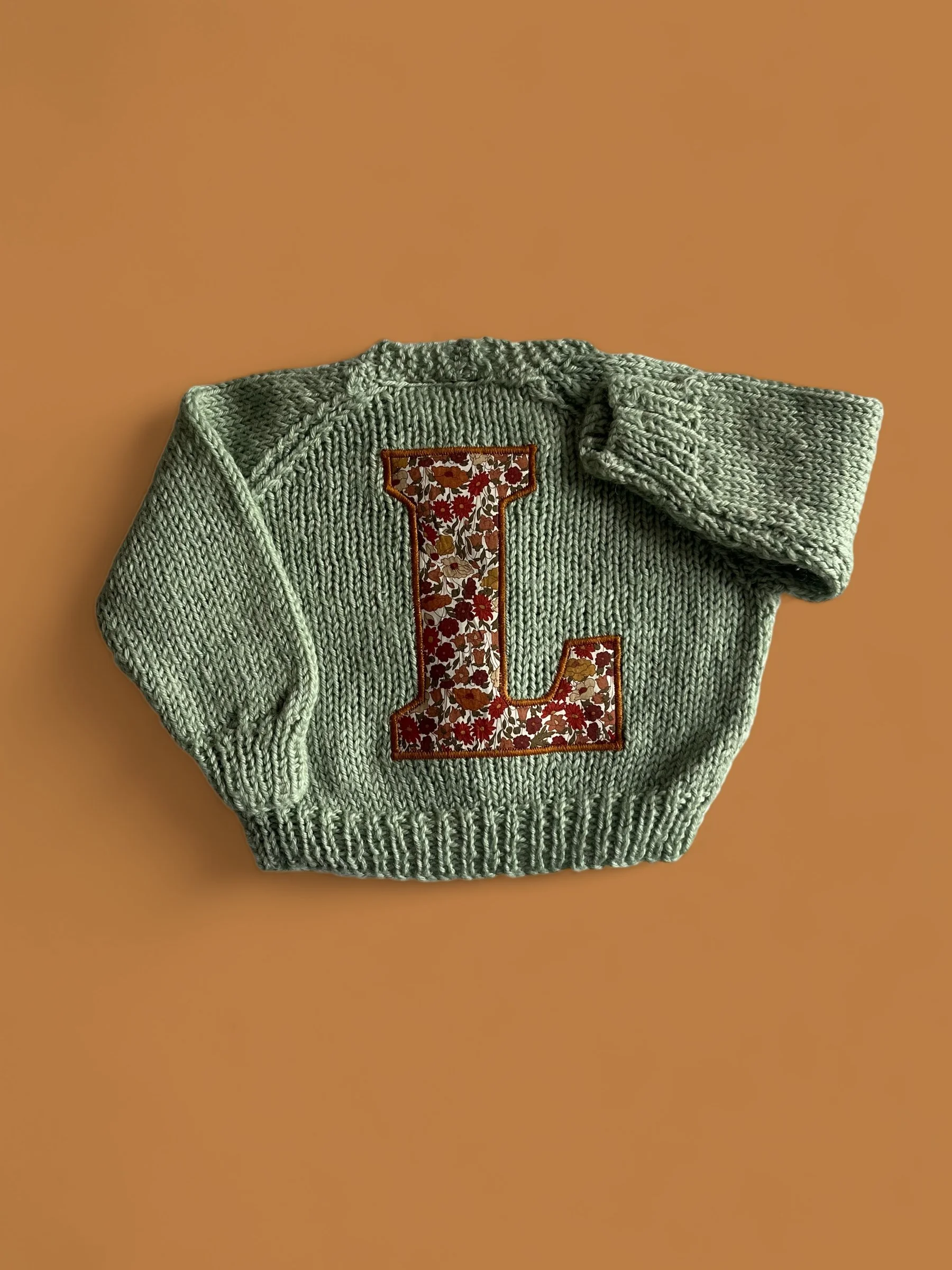 A green knitted sweater with a large letter 'L' patch featuring a floral pattern, laid flat on a brown background.