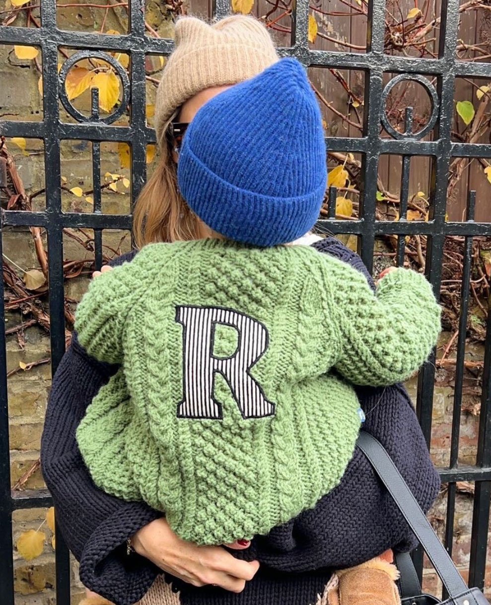 Person holding a child wearing a green sweater with the letter R on the back. The child also wears a blue knit beanie. The adult is wearing a beige hat and stands in front of a wrought iron fence with vines.