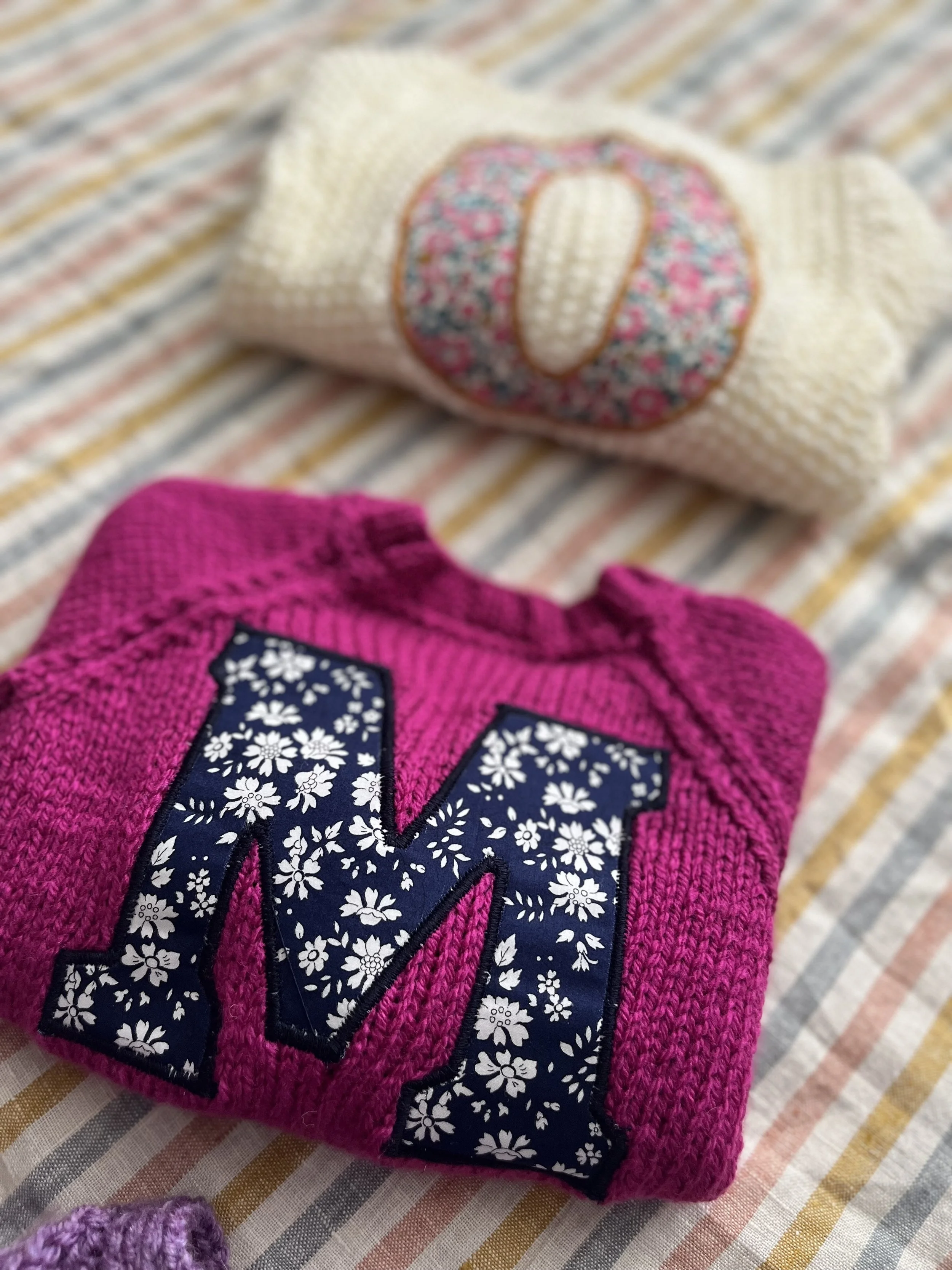 Two folded sweaters on a striped fabric background, one white with a floral "O" and one purple with a floral "M".