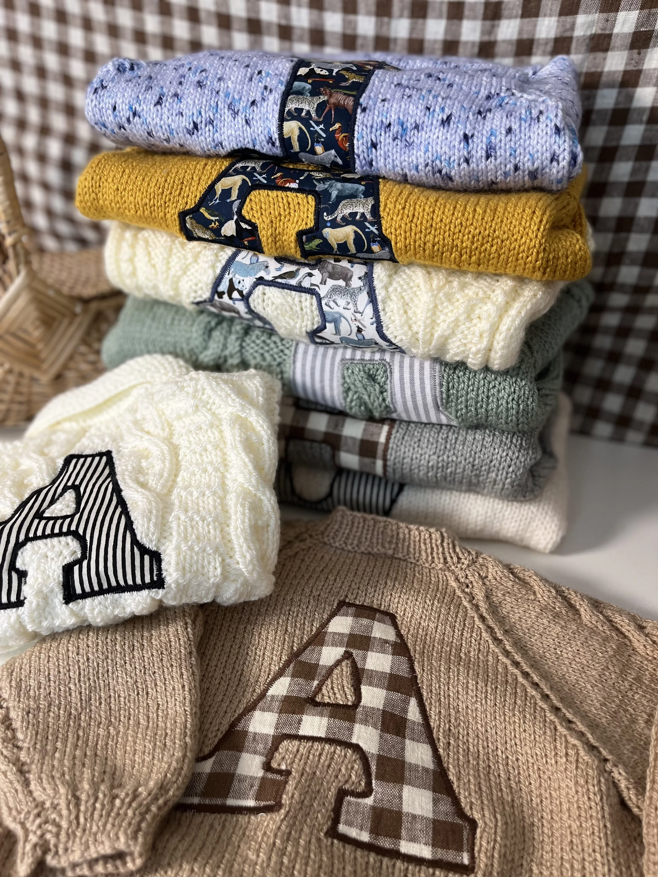 Stack of folded sweaters with large letter 'A' patches on each, in various colors and patterns, arranged on a checkered surface.