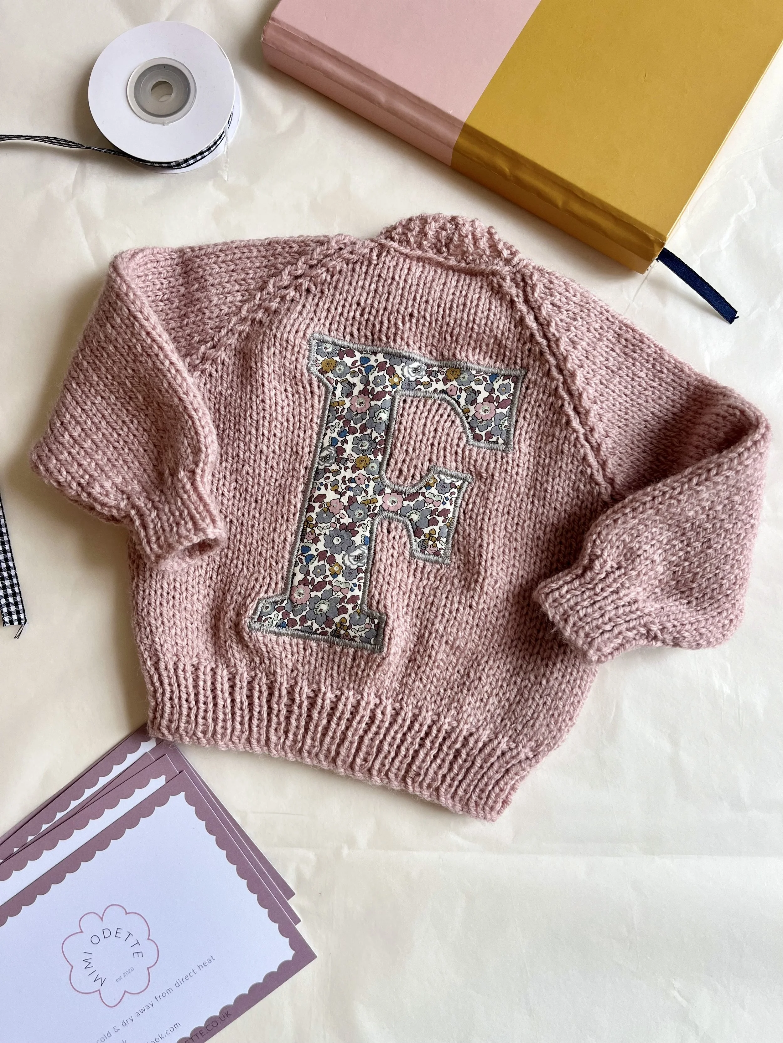 Pink knitted baby sweater with floral letter F, surrounded by gift packaging materials and a card.