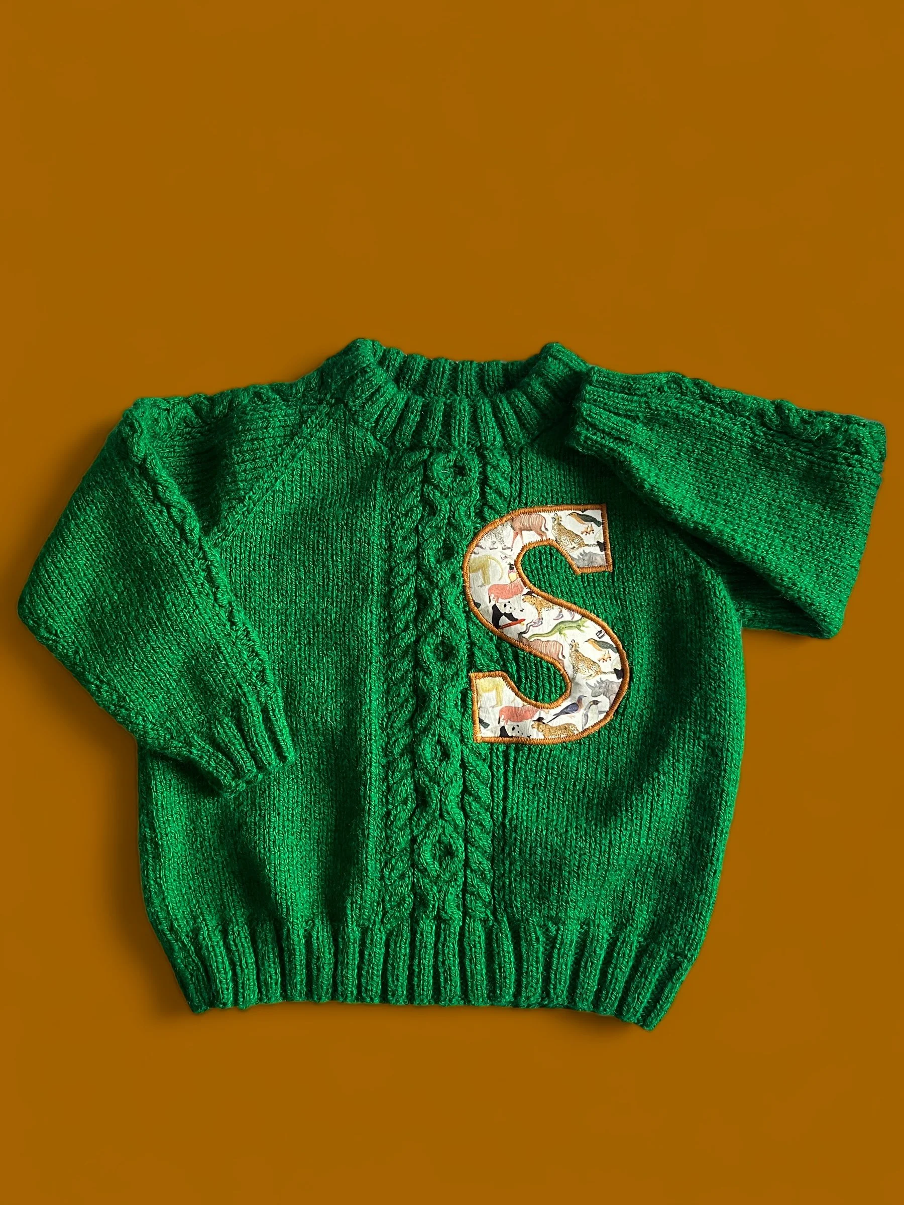 Green knitted child's sweater with a large embroidered letter 'S' on the front, adorned with various animal illustrations, placed on a brown background.