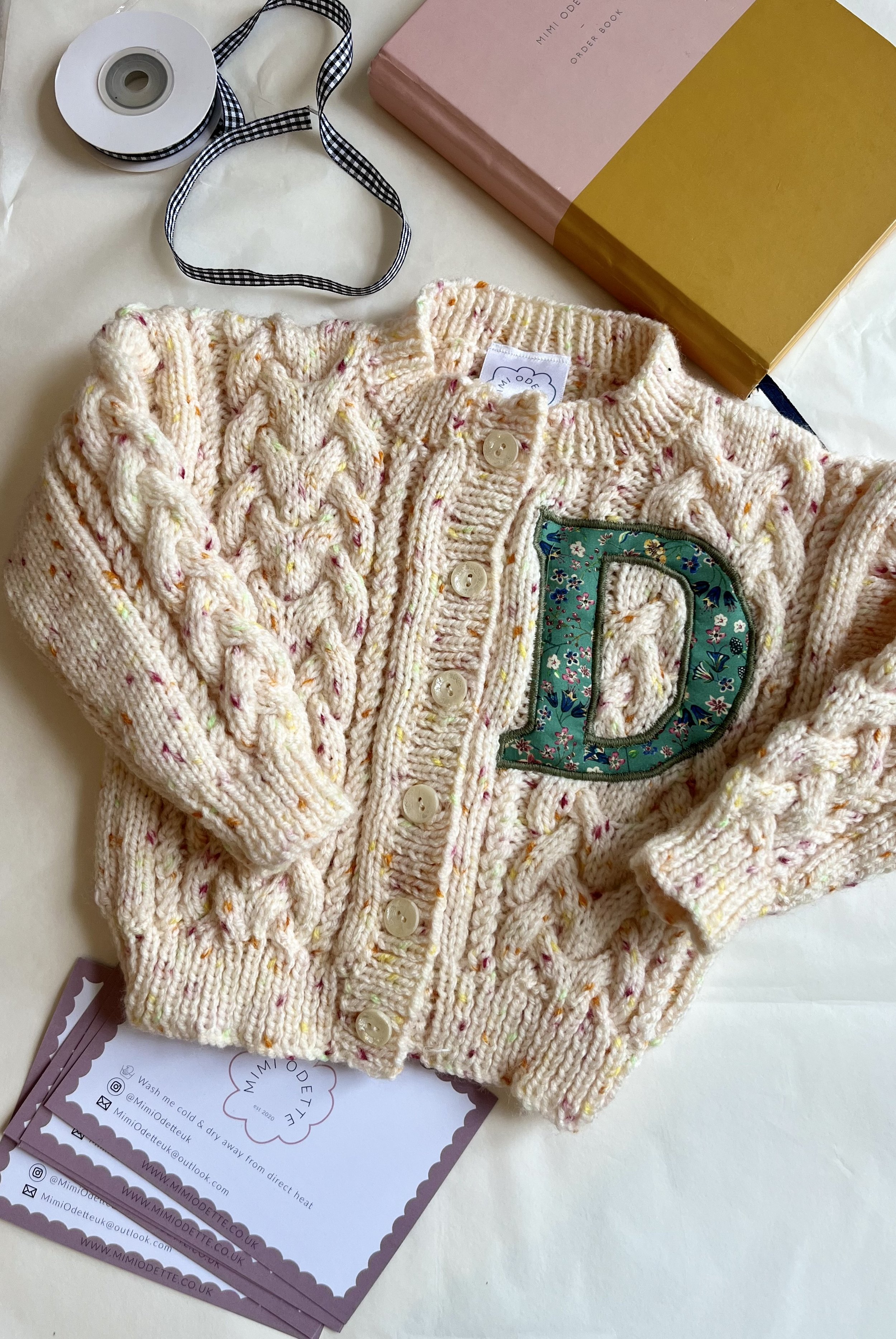 Knitted baby sweater with a letter "D" patch, surrounded by a ribbon, a book, and business cards.