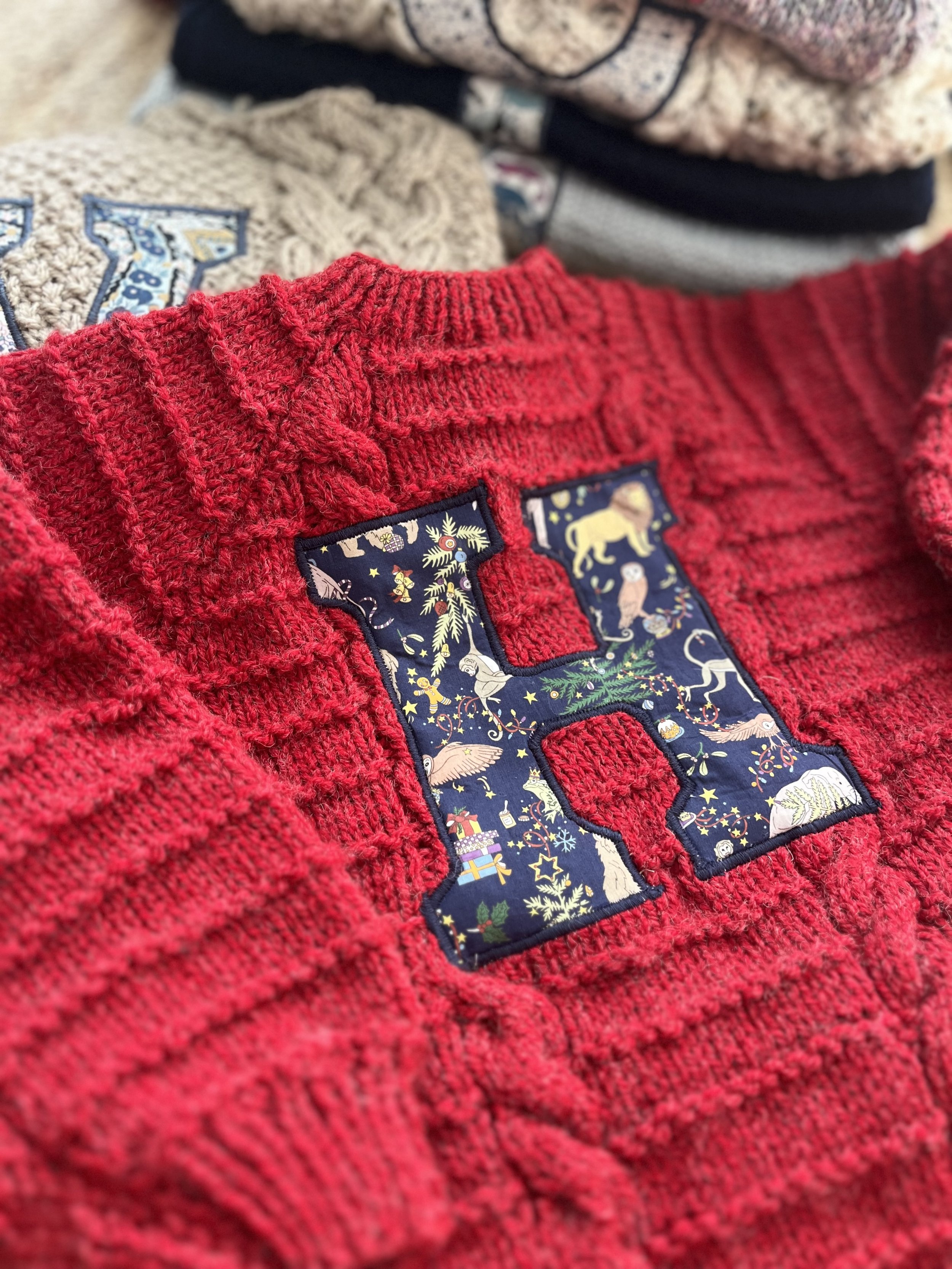 Close-up of a red knitted sweater with a large letter 'H' decorated with intricate patterns, lying on top of other folded sweaters.