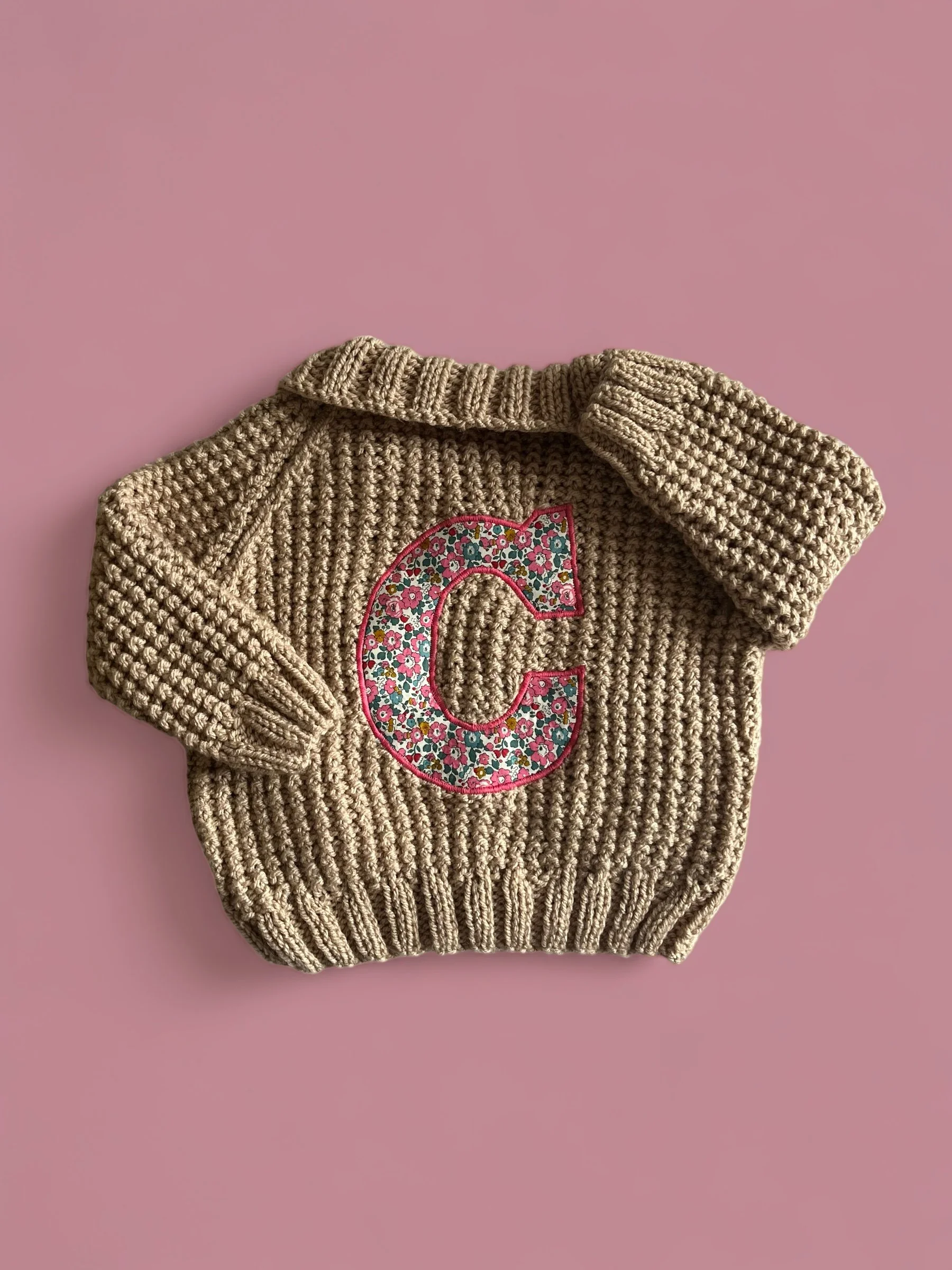 Knitted beige sweater with a floral-patterned letter C on a pink background.