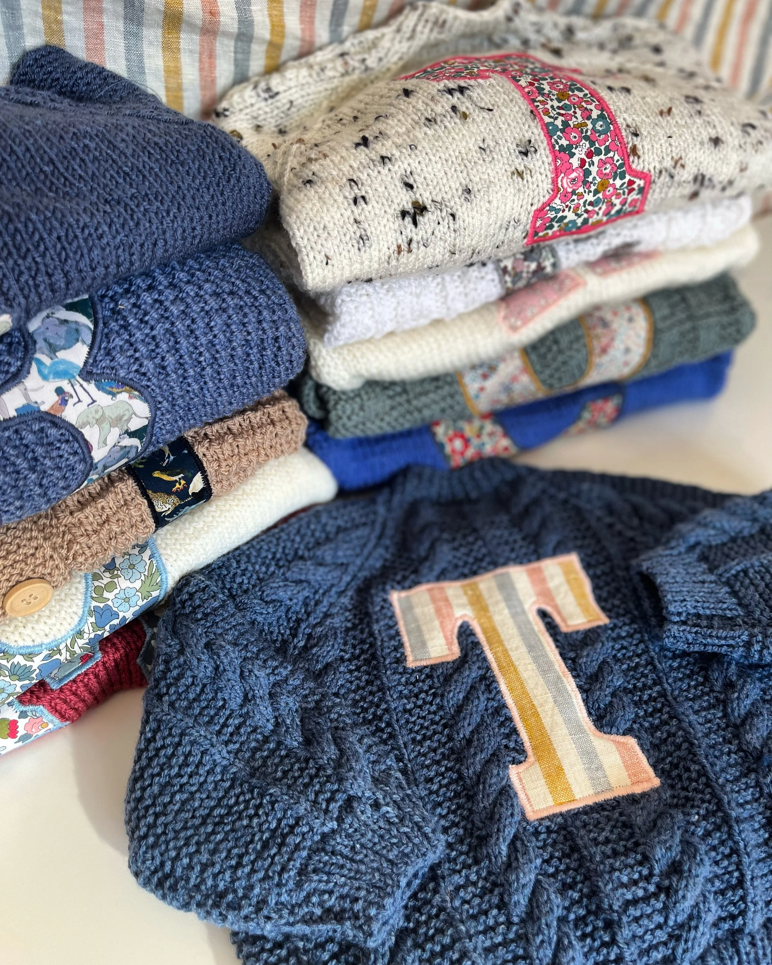 A stack of colorful knitted sweaters embellished with patterned patches featuring different letters and designs, arranged on a striped fabric background.