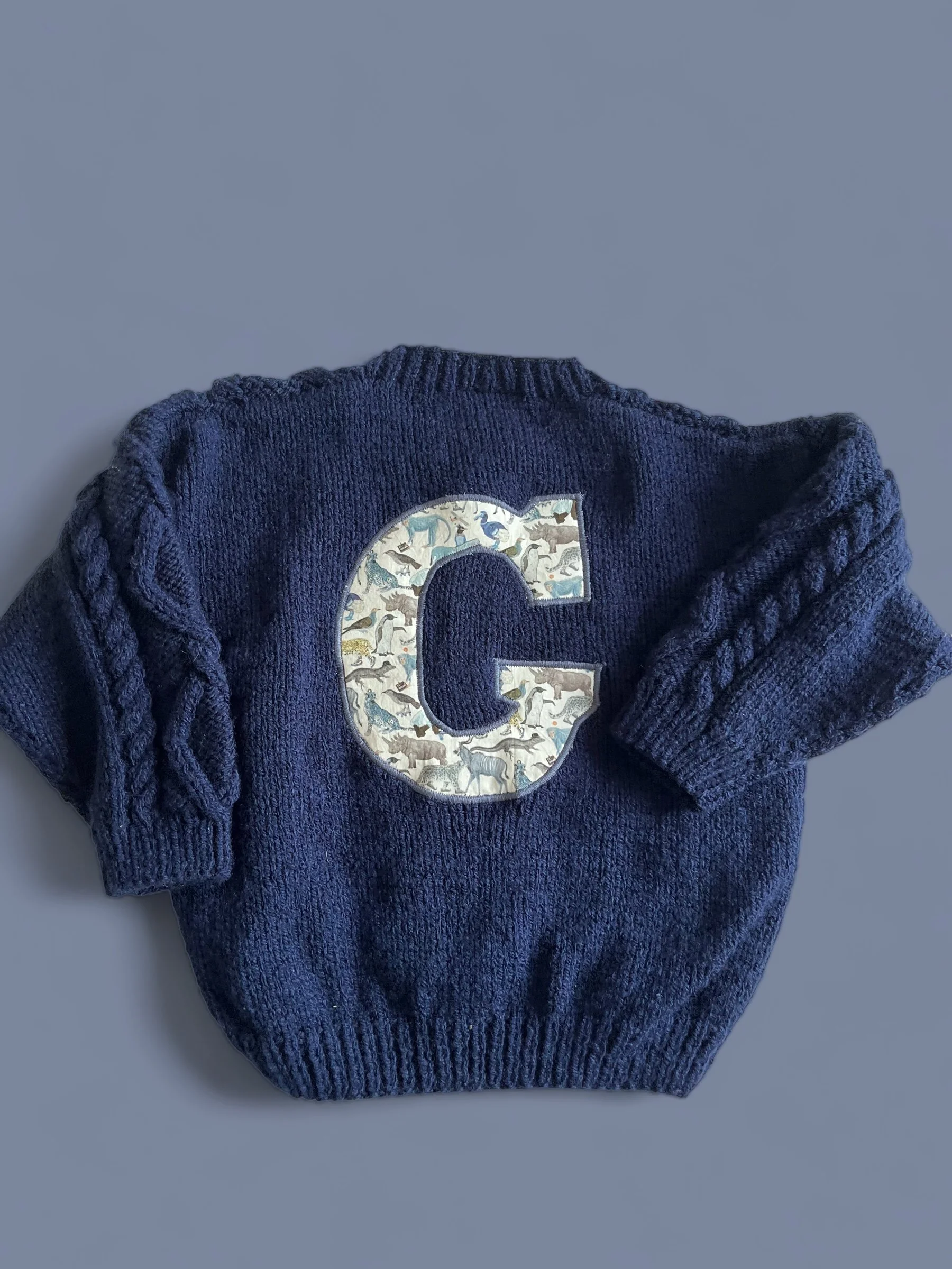 A navy blue knitted sweater with a large letter 'C' patch on the front, featuring a pattern of various animals.