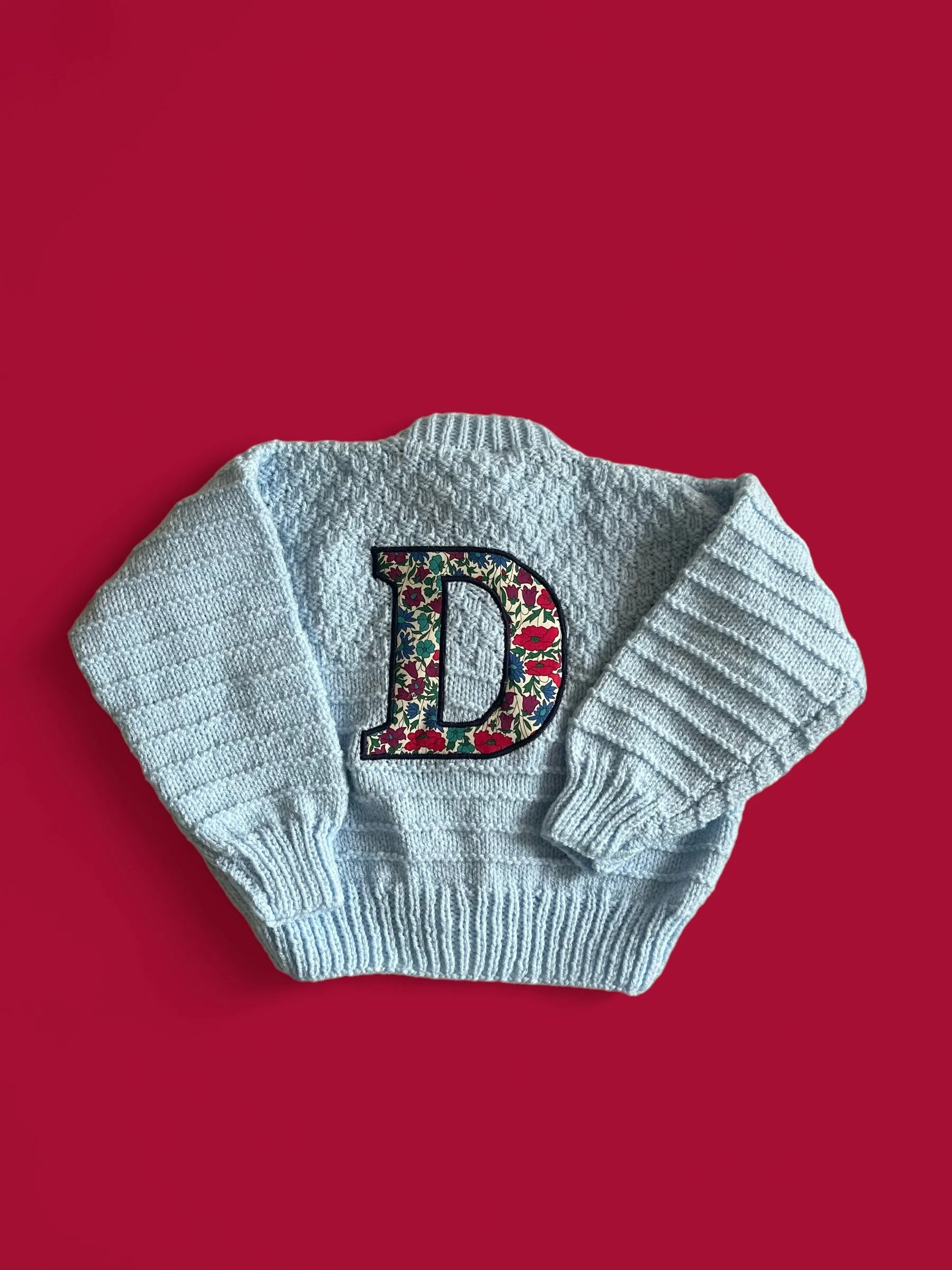 A gray knitted sweater with a floral embroidered letter D on the front, laid flat on a red background.