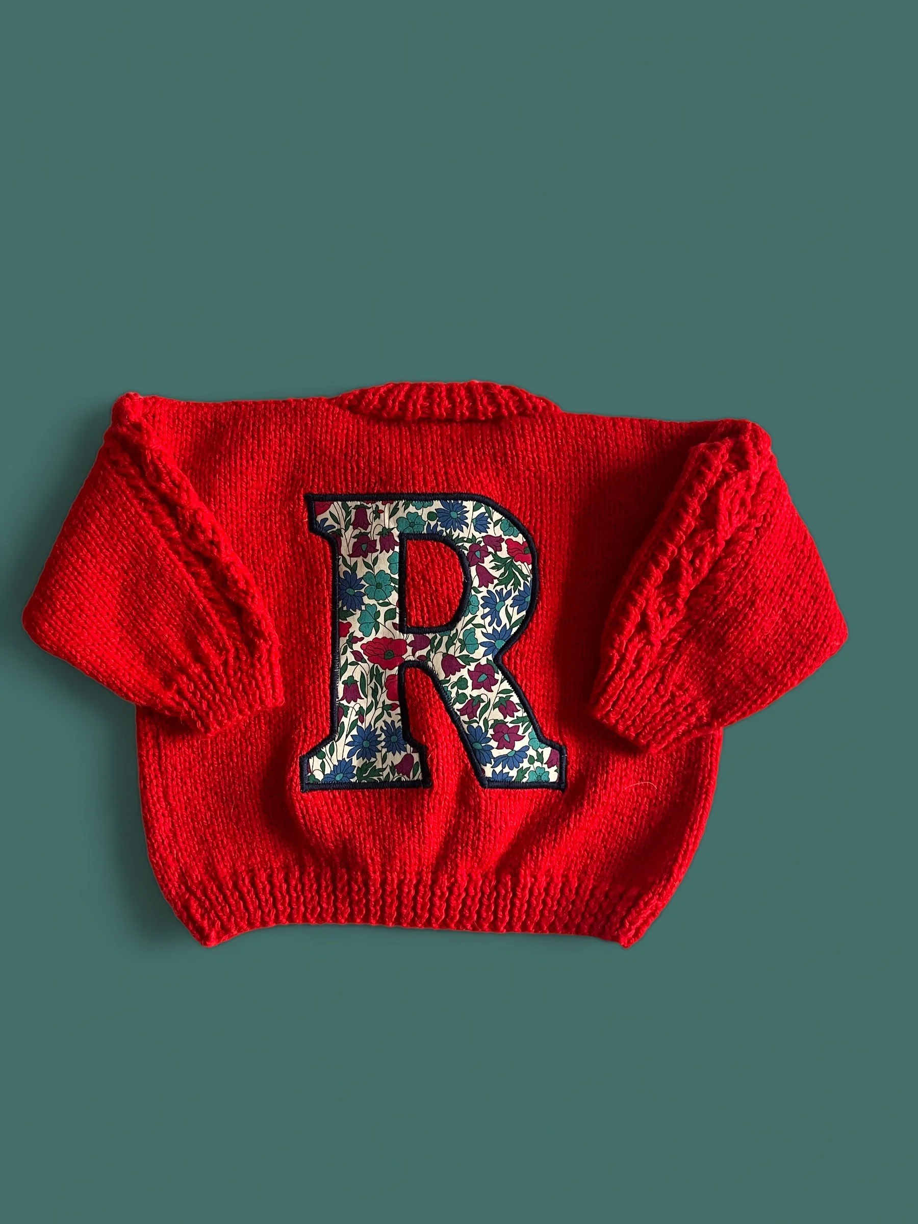 Red knitted child's sweater with a large floral letter R appliqué in the center.