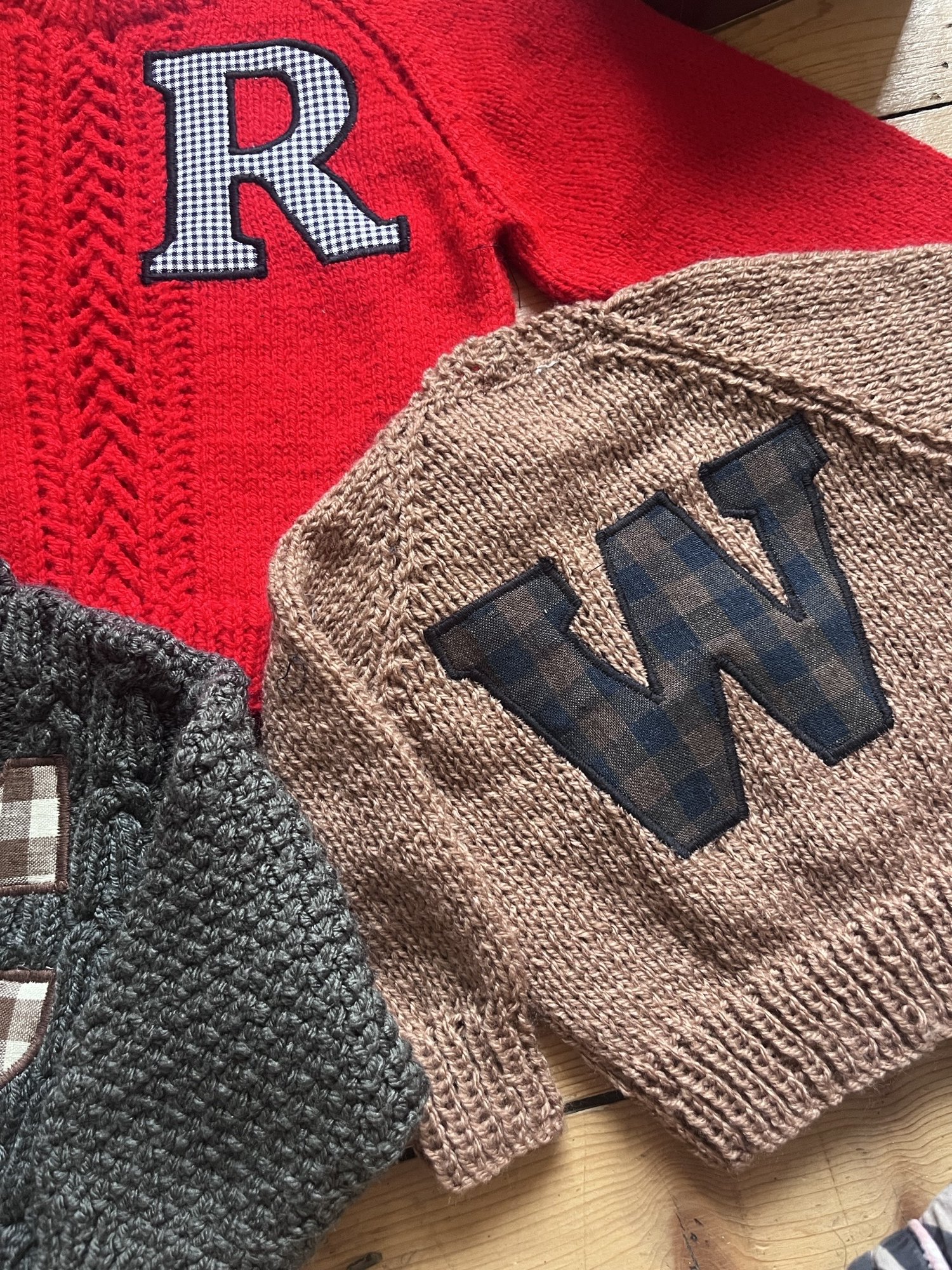 Close-up of three knit sweaters with letter patches: a red sweater with a white and black checkered 'R', a beige sweater with a dark checkered 'W', and a gray sweater with a checkered emblem.