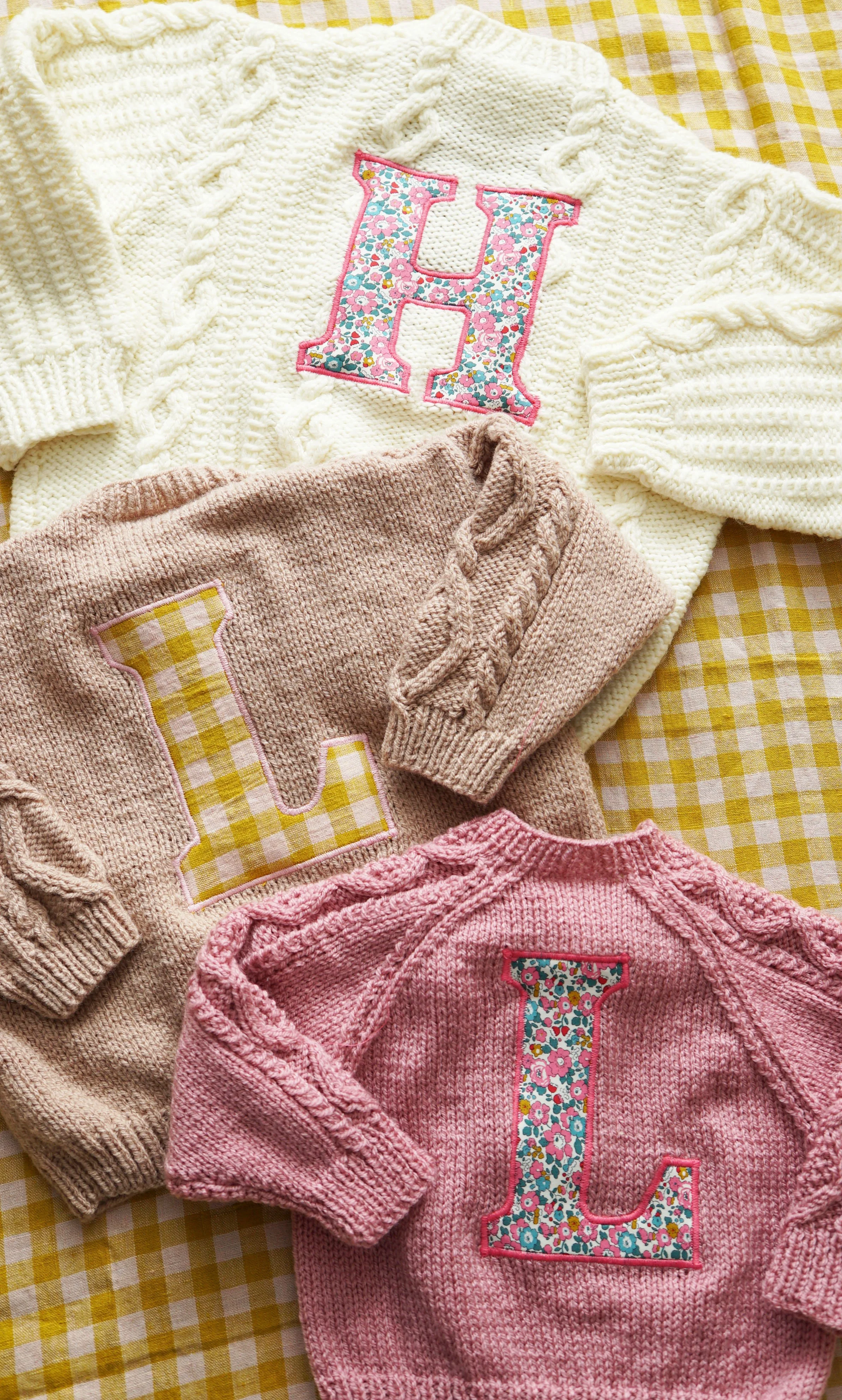 Three knitted sweaters on a yellow gingham background, each with a large letter appliqué: a white sweater with an 'H' in floral fabric, a beige sweater with an 'L' in checker pattern, and a pink sweater with an 'L' in floral fabric.