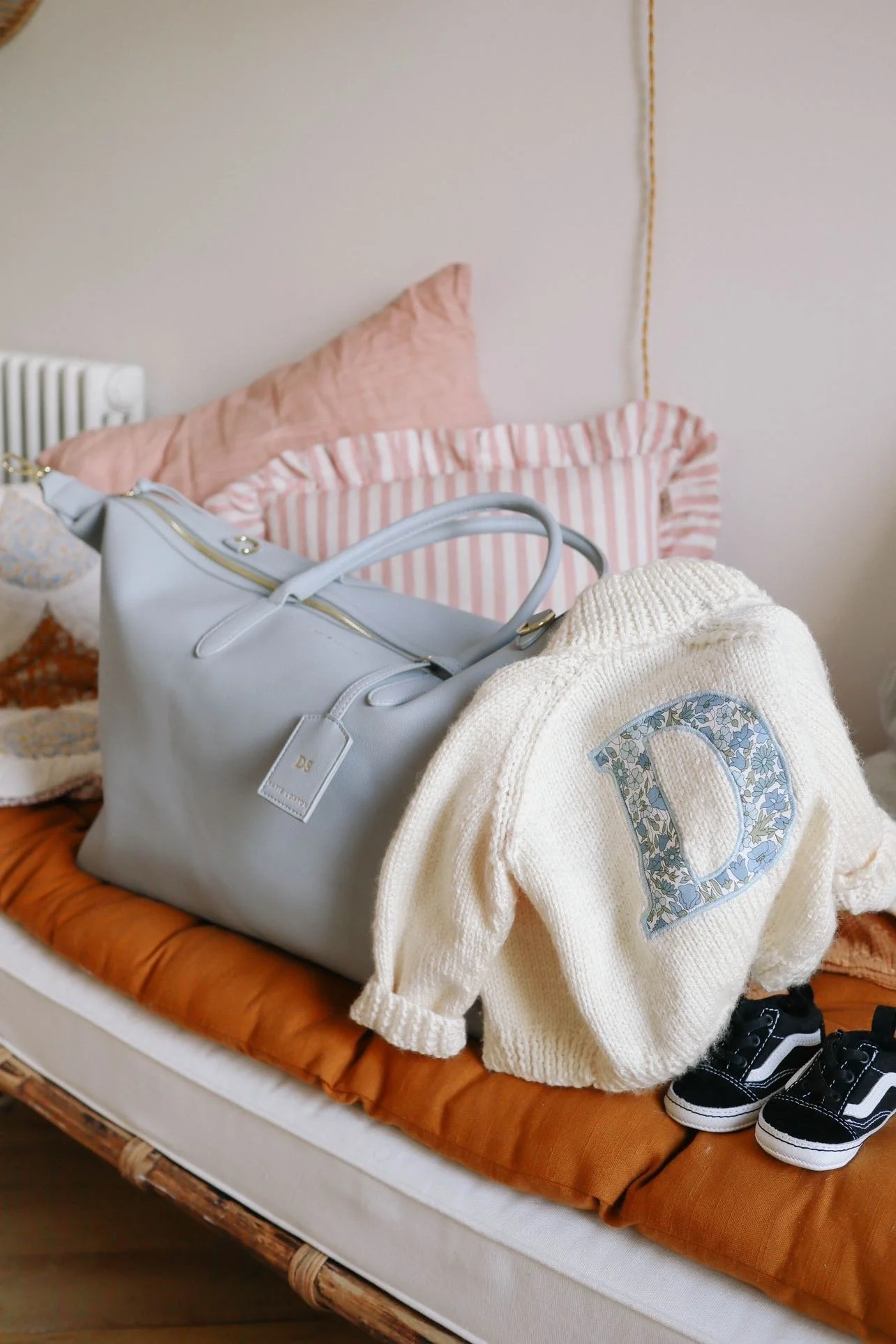 A bed with pink and striped pillows, a white handbag, a cream-colored sweater with a floral 'D' on it, and black sneakers placed on a brown bedspread.