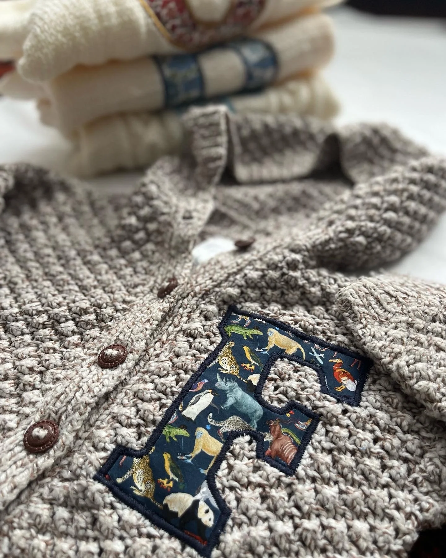Close-up of a knitted beige sweater with brown buttons and an embroidered letter 'F' decorated with various animals, resting near a folded cream blanket with a patterned edge.