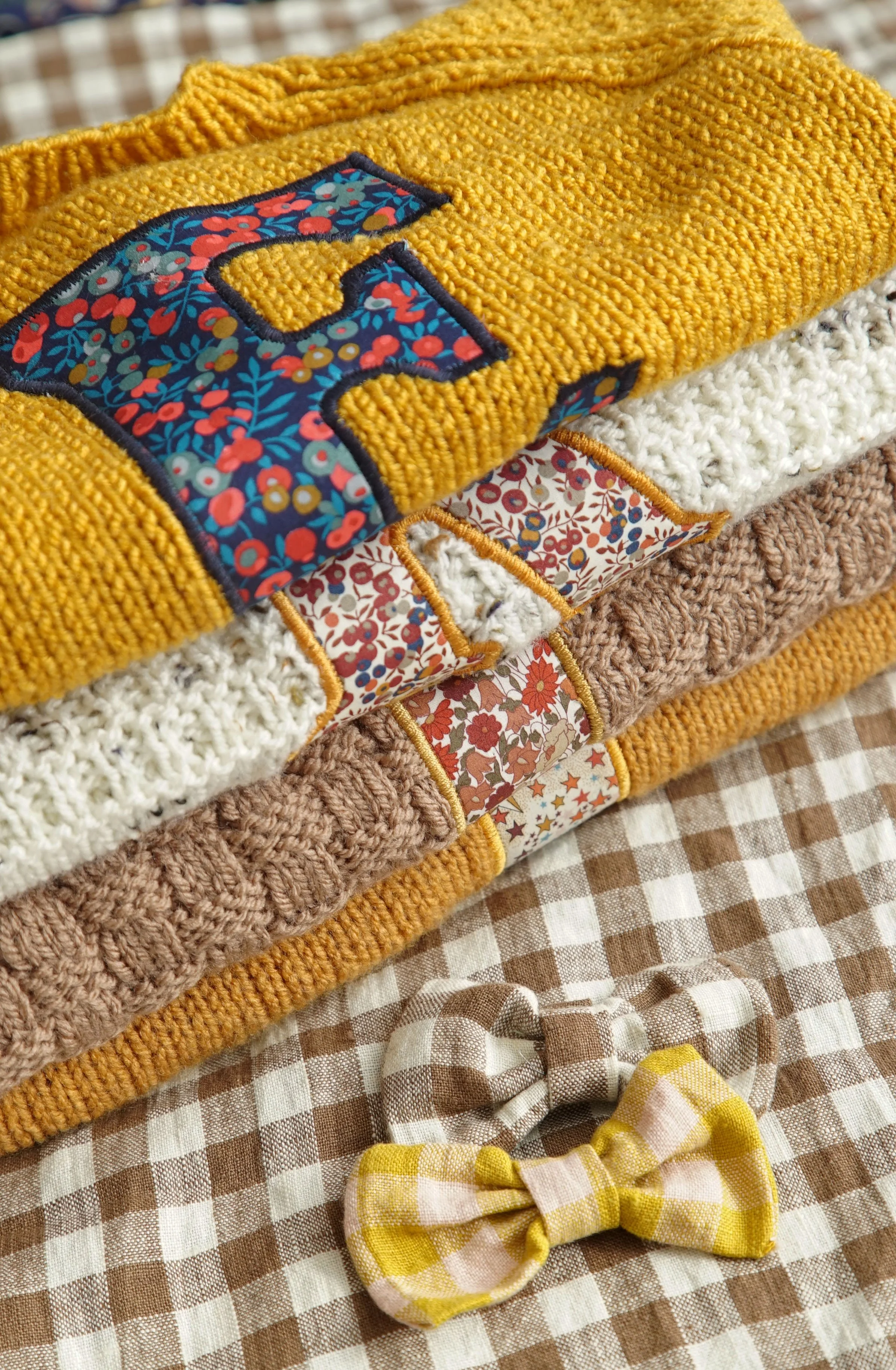 Folded knitted sweaters with floral patterns, alphabet applique, and two plaid bows on a checkered fabric.