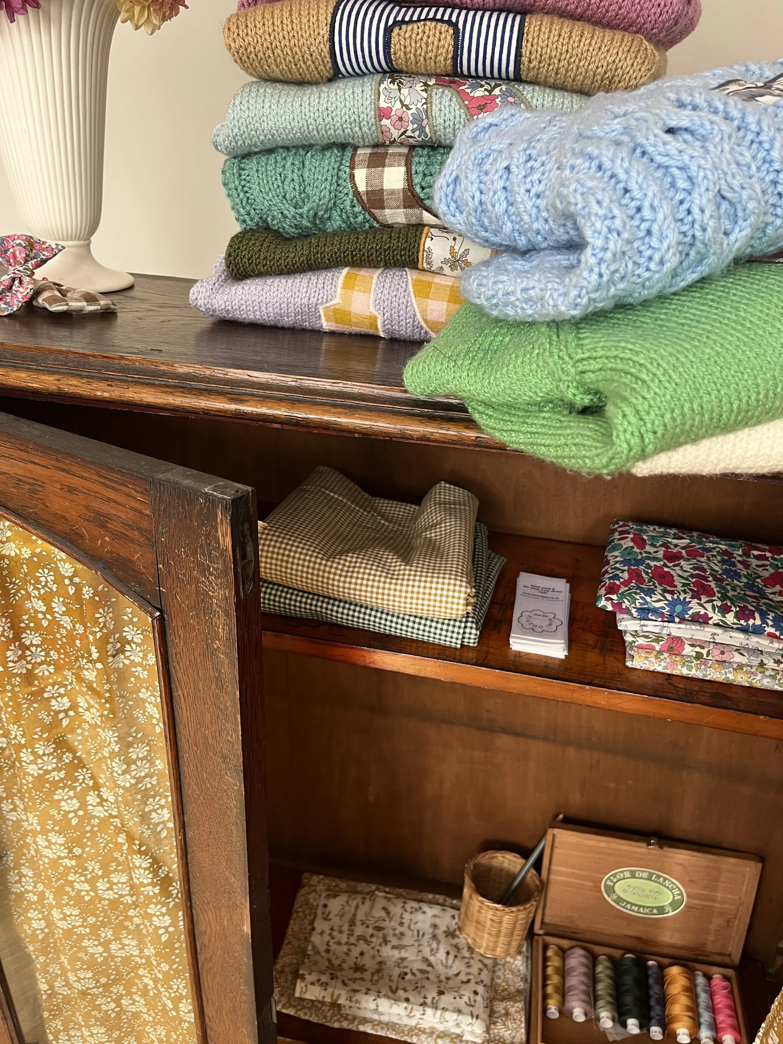 Stacked knitted and fabric items on a wooden cabinet with sewing accessories.