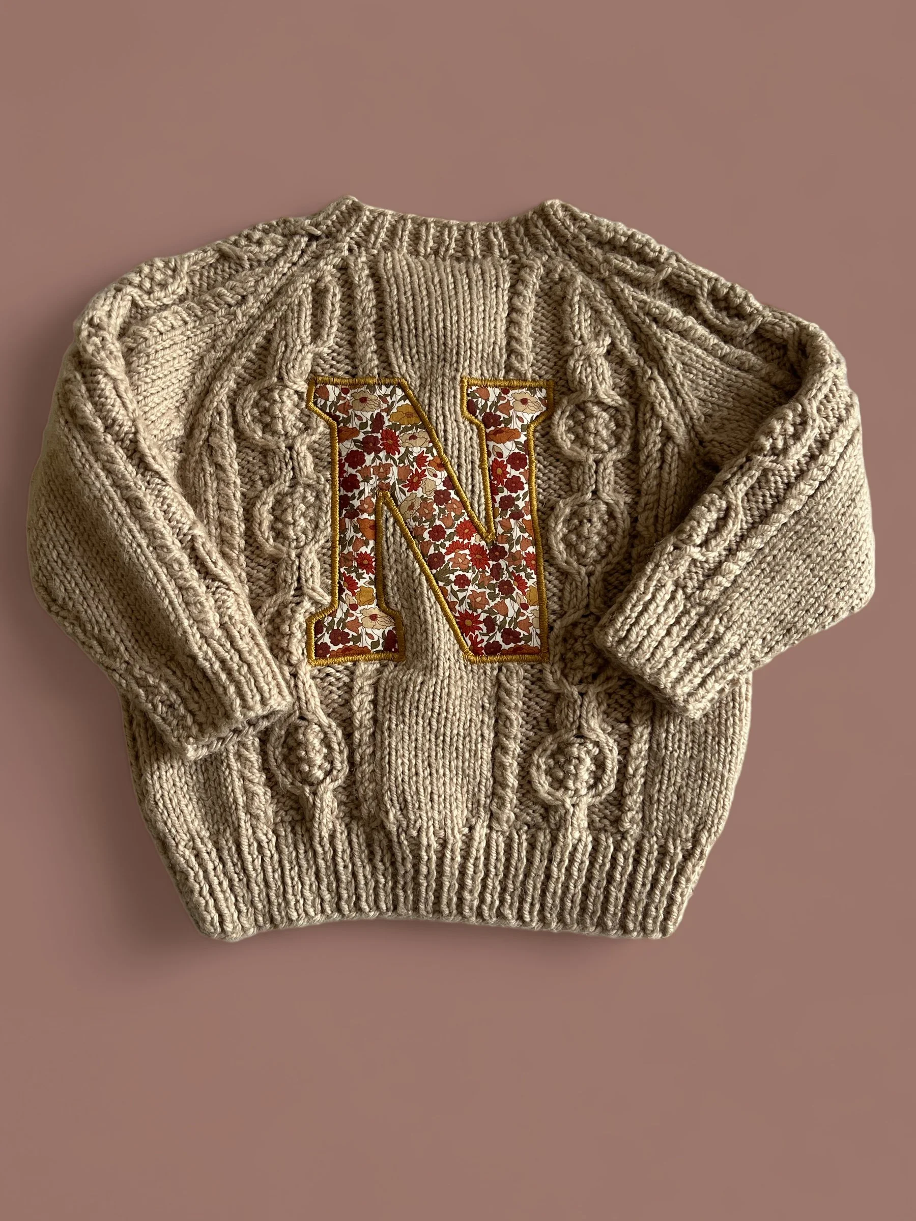 Cream-colored knit sweater with a large embroidered letter 'N' filled with a floral pattern, placed on a pink surface.