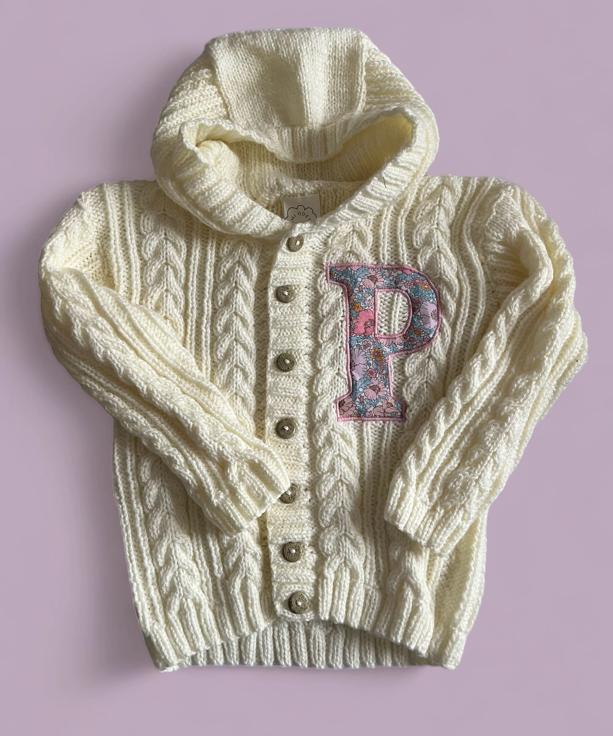 A cream-colored knitted hoodie with cable knit patterns and a floral embroidered letter 'P' patch on the front.