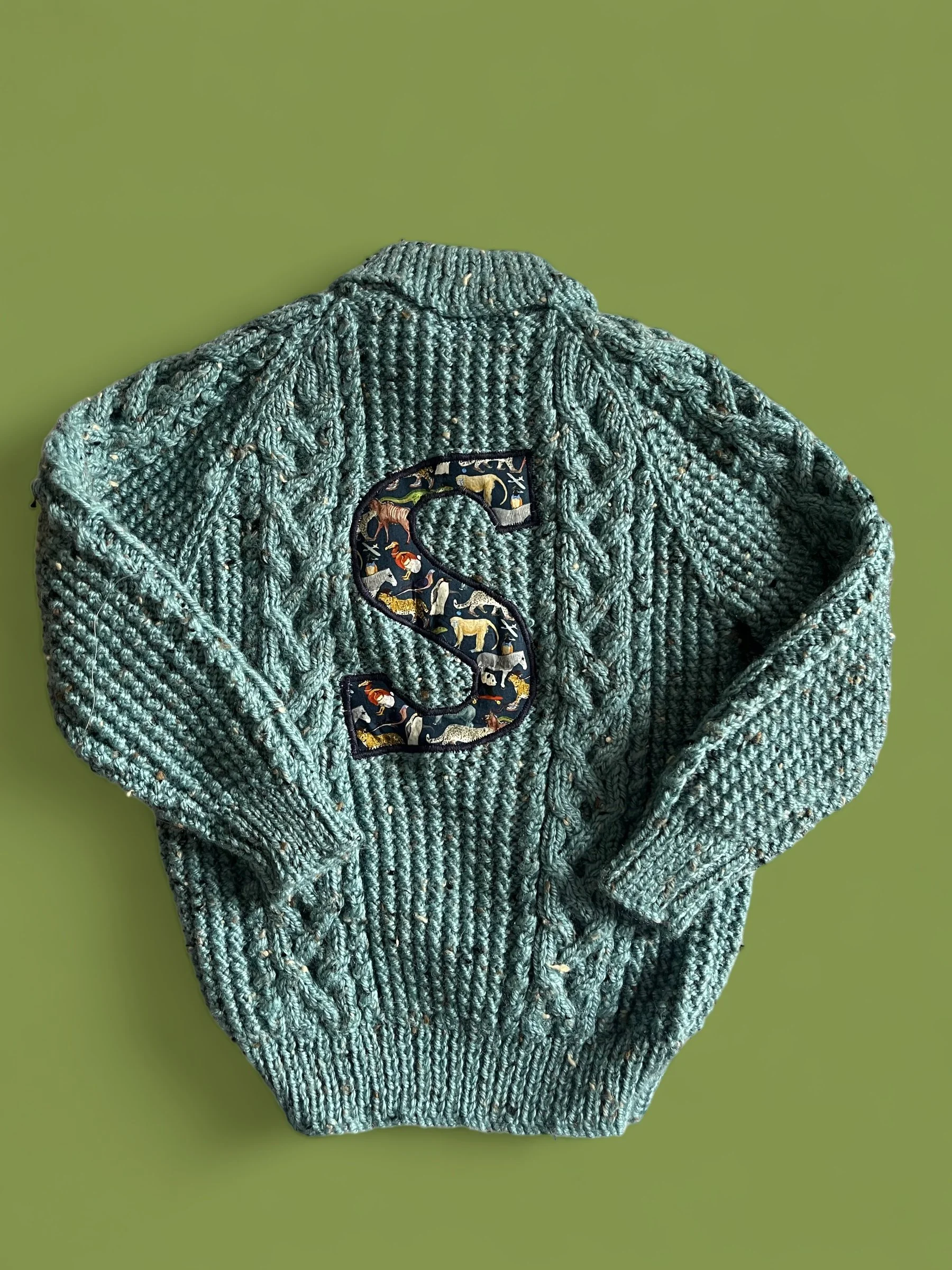 A knitted sweater with a large embroidered letter 'S' on the front, featuring a colorful animal pattern, laid flat on a green surface.
