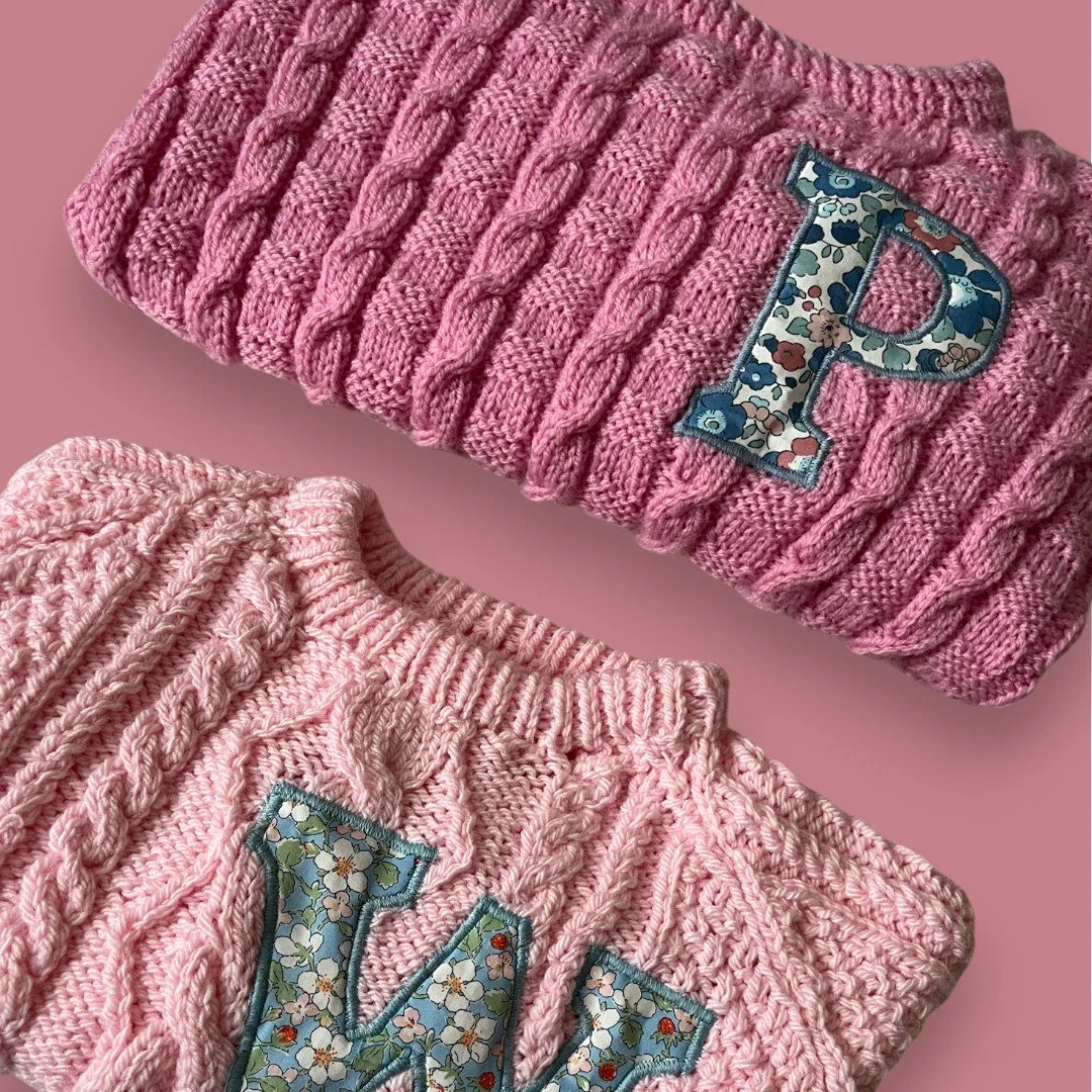Two pink knit sweaters with embroidered floral letters, one with a large 