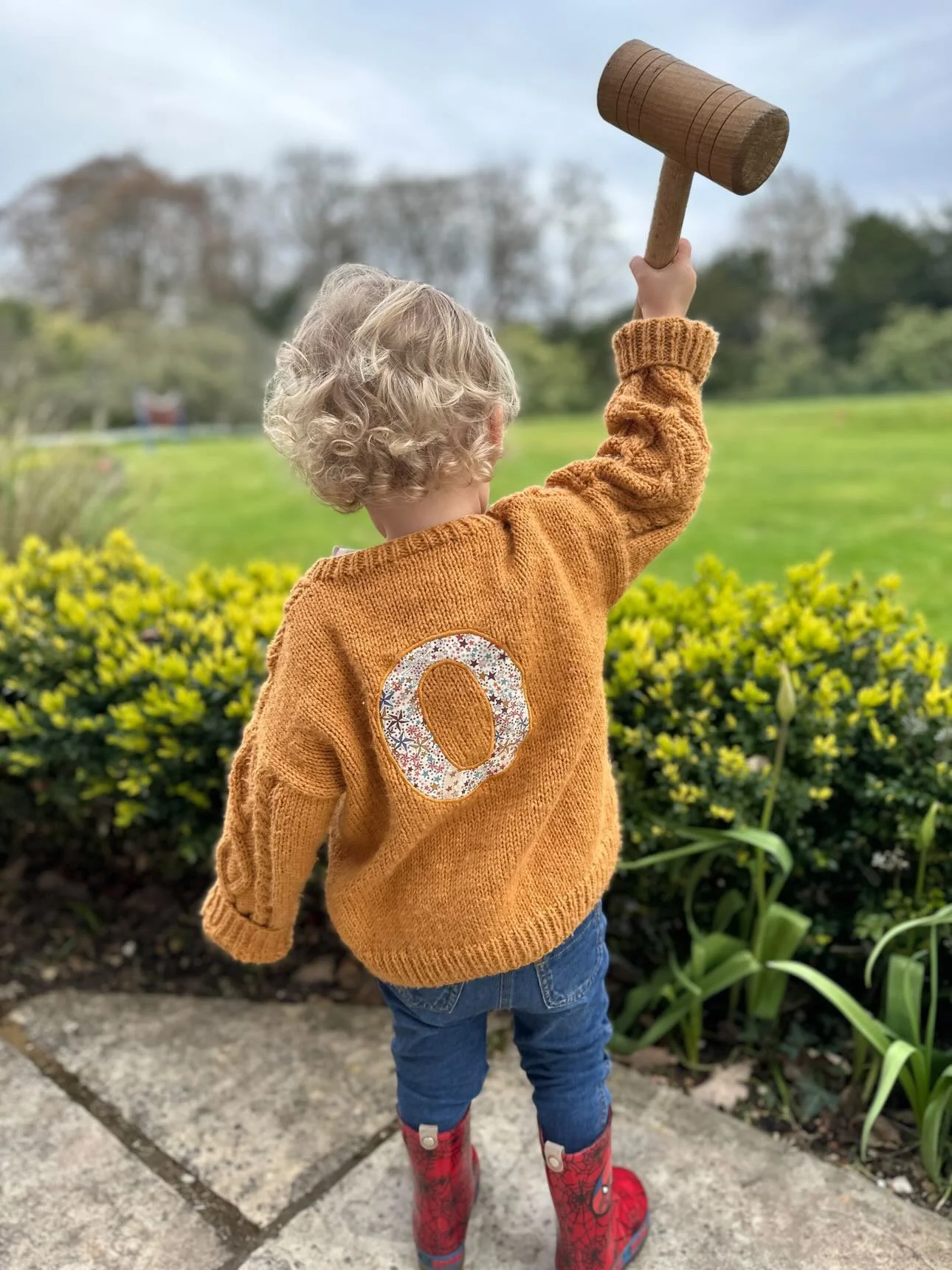 A child with curly blonde hair wearing an orange sweater with the letter O on the back, holding a toy wooden mallet, standing outside on a stone path in front of bushes and a grassy area.