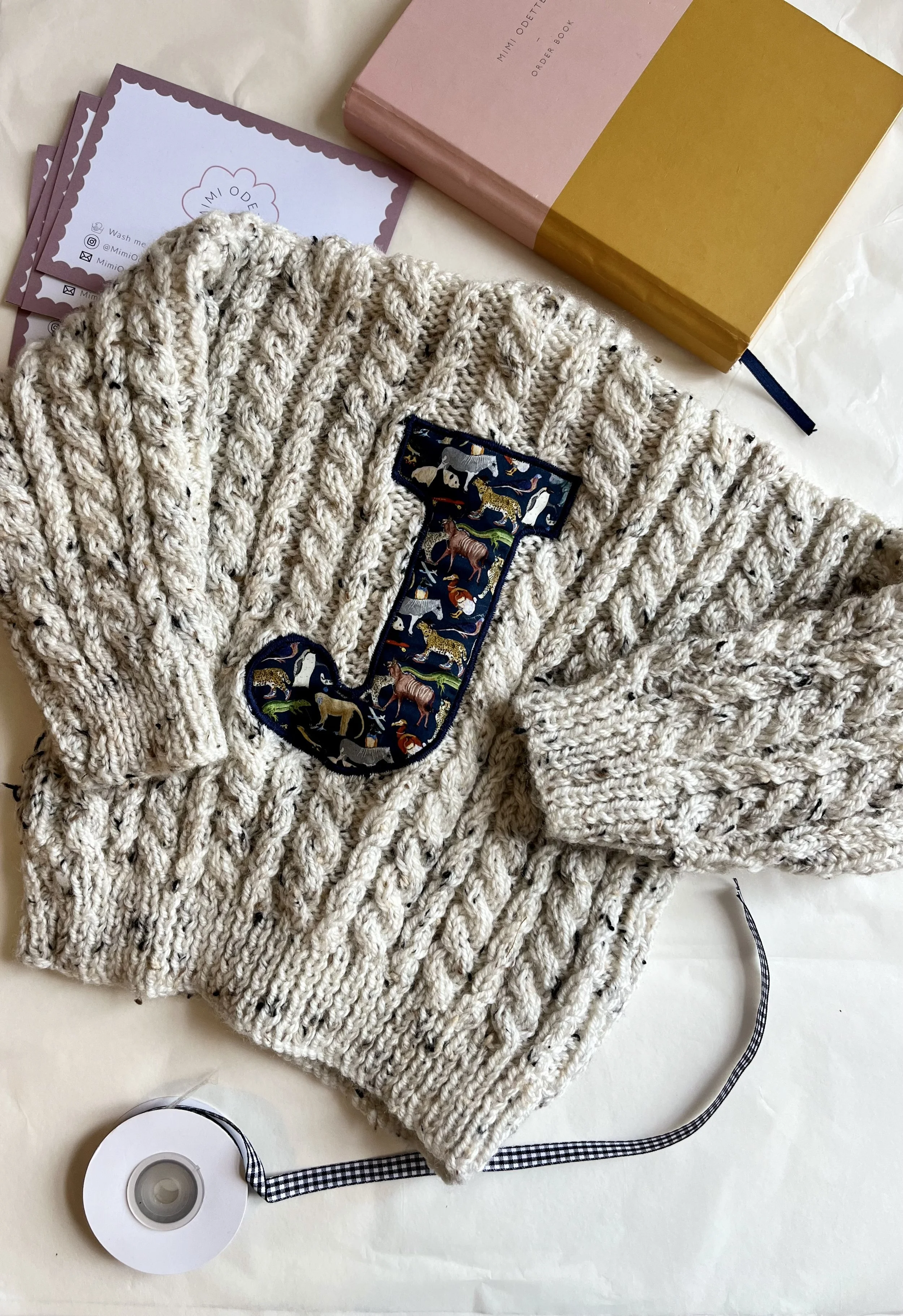 Knitted sweater with a large 'J' applique, surrounded by a pink and gold book, cards, and a roll of black-and-white checkered ribbon on a white background.