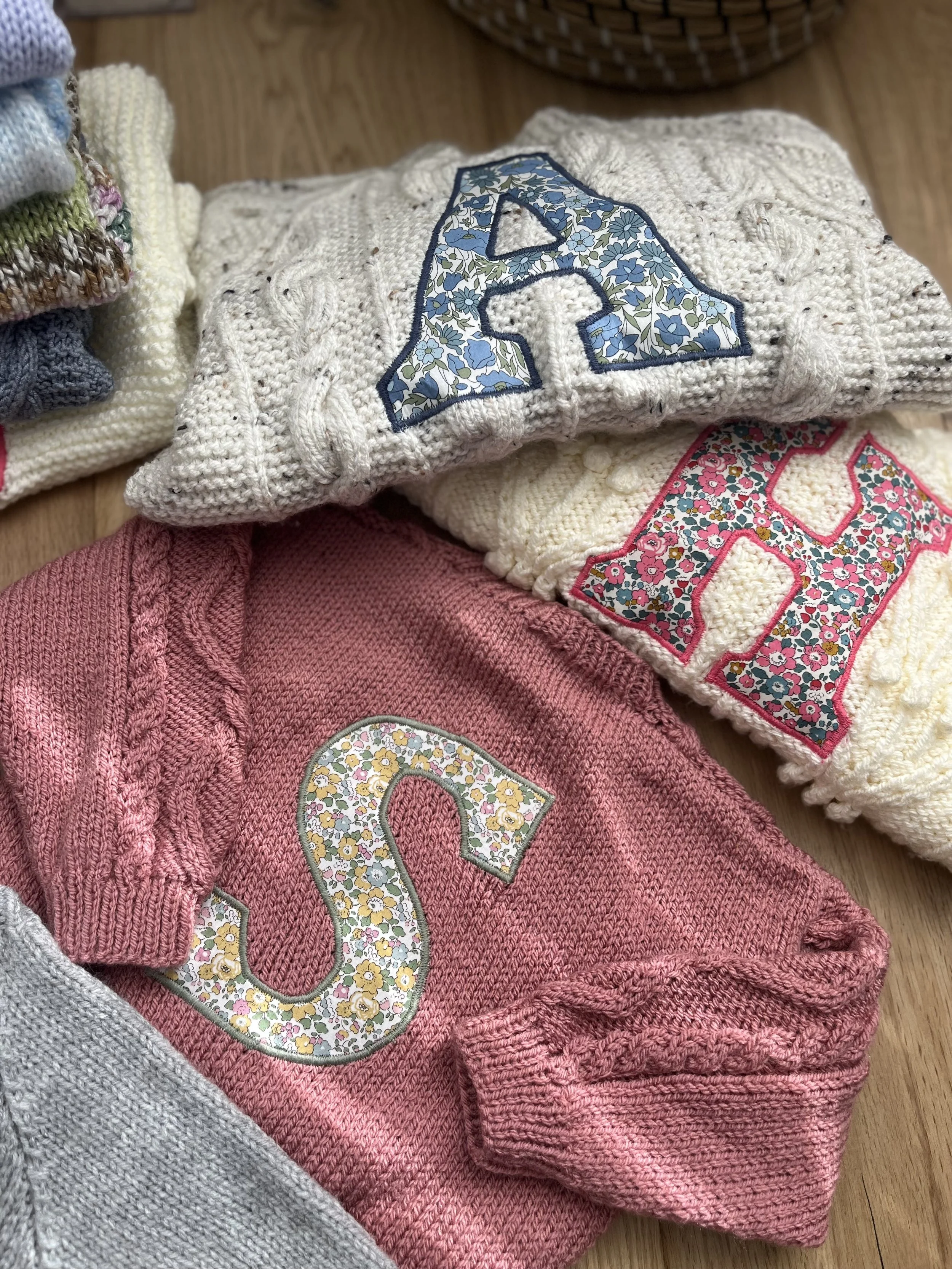 Knitted sweaters with floral letter appliques in various colors, folded on a wooden surface.