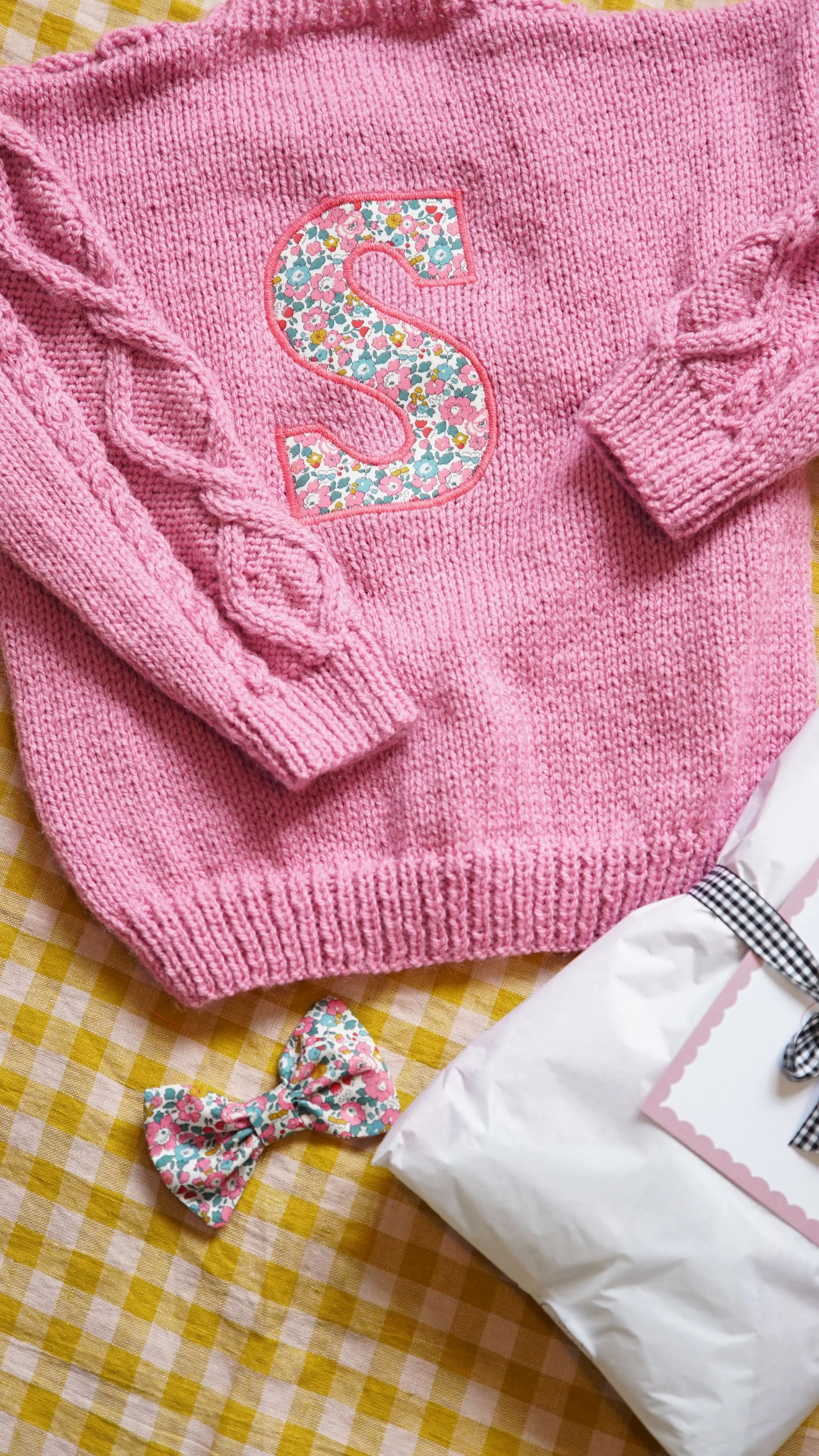 Pink knitted sweater with floral "S" patch, floral bow, and wrapped package on yellow plaid fabric.