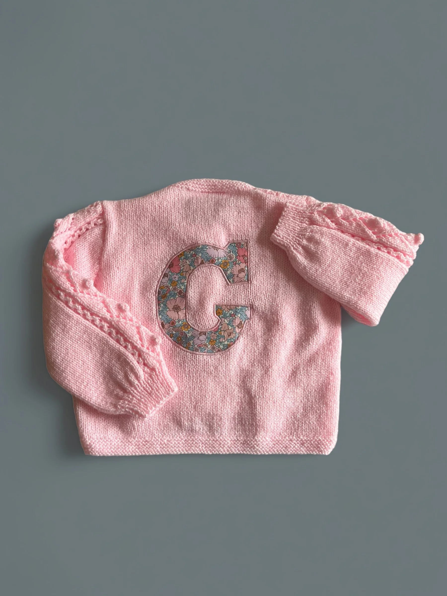 Pink baby sweater with a floral-patterned letter 'C' on the front, laid flat on a gray surface.