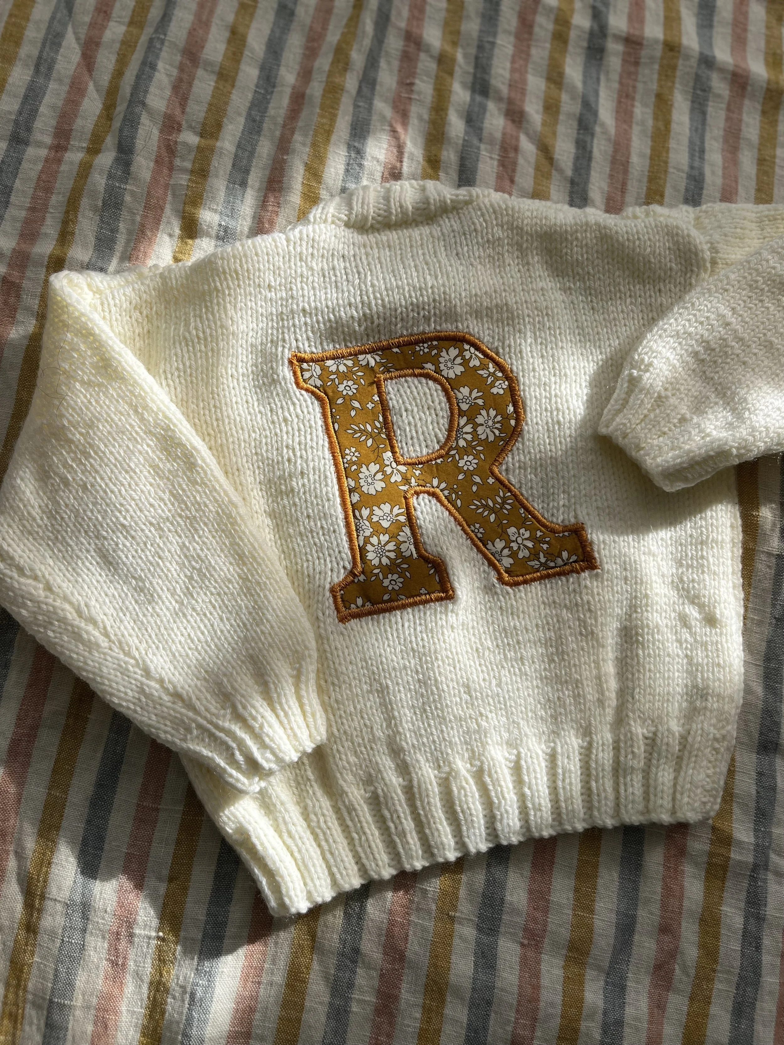 White knitted sweater with a large floral-patterned letter 'R' on striped fabric background.
